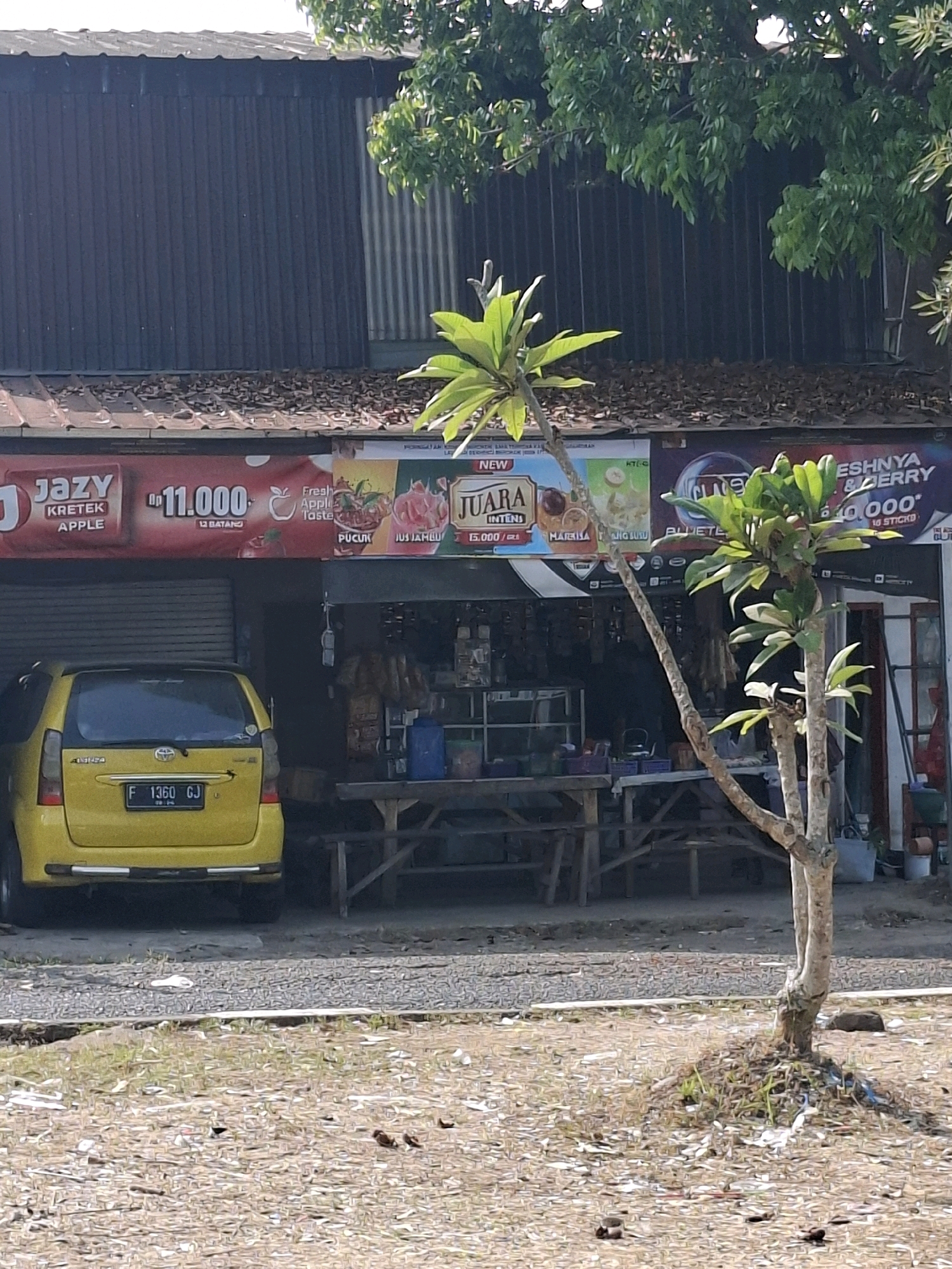 The image captures a street scene in Sukabumi, Indonesia. A yellow Toyota Avanza with license plate F 1360 GJ is parked in front of a row of shops. The shops have awnings with advertisements for various products, including "Jazy Kretek Apple" and "Juara Intensi." One of the shops appears to be a convenience store or warung, with shelves stocked with goods visible through the open entrance. In the foreground, a young tree with sparse leaves stands on a patch of dry ground scattered with debris. The background shows a building with a corrugated metal roof and a dark-colored facade, likely part of the shops. Lush green foliage from trees overhangs the scene from above. The overall impression is of a typical urban commercial area in Indonesia.