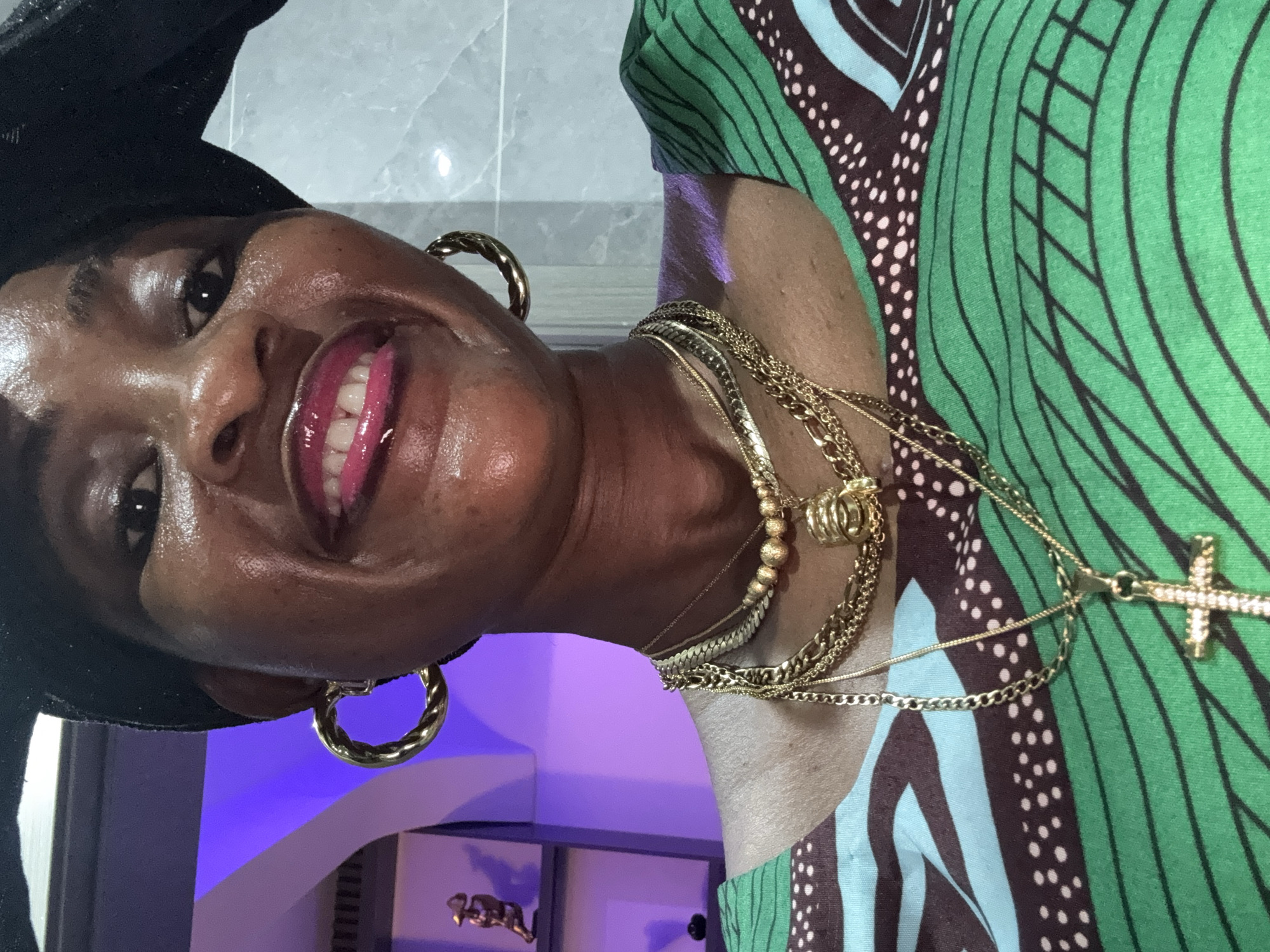 A close-up, selfie-style photograph shows a Black woman from the chest up, wearing a green and brown patterned dress. She has on a black headwrap, large gold hoop earrings, and multiple gold necklaces. One necklace has a cross pendant. Her lips are painted a bright pink, and she is smiling. She appears to be indoors, with a purple-lit shelf visible in the background on the left, and a light-colored tiled wall on the right. There is no indication of the specific location within Ajara, Nigeria.