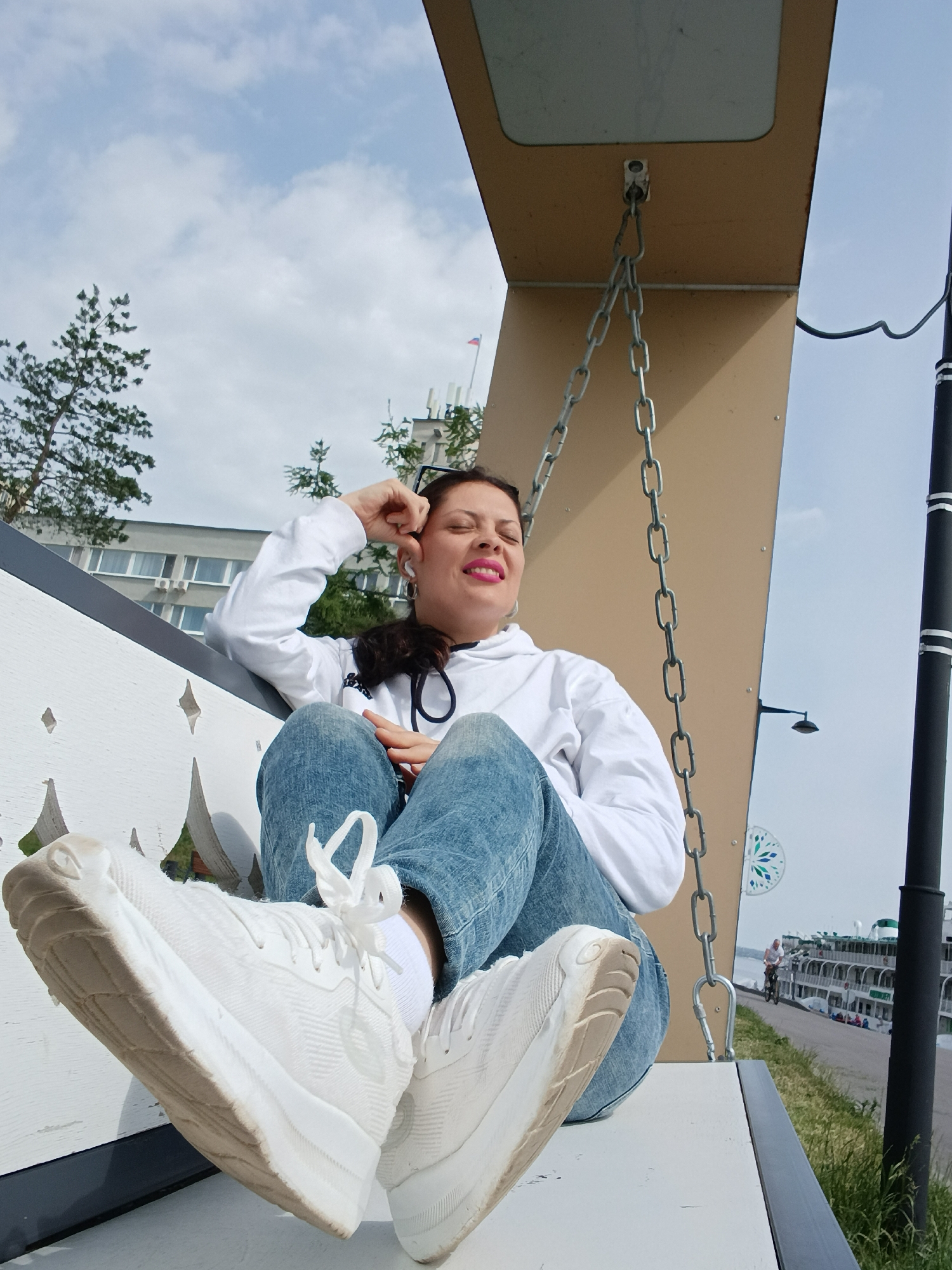 A woman is seated on a white swing bench, captured from a low angle. She wears a white hoodie, blue jeans, and white sneakers, with her feet propped up on the swing. Her dark hair is partially visible, and she has bright pink lipstick. White earbuds are in her ears, and she appears relaxed, with her eyes closed or squinted, and one hand resting near her forehead. Her expression suggests she might be enjoying music, the moment, or reacting to the sunlight.
The swing is suspended by metal chains from a light brown, angular structure. In the background, green trees are visible against a bright, cloudy sky. A multi-story building with numerous windows can be seen through the trees. Further back, a large multi-deck cruise ship is docked alongside what appears to be a paved waterfront promenade or path. A small figure on a bicycle is also visible on this path near the ship. A black pole, possibly a lamppost, stands on the right side of the frame. The overall scene suggests a pleasant daytime setting, likely a park or embankment area by a body of water. This environment is consistent with the city of Saratov, Russia, which is located on the Volga River. No clear, readable text is visible in the image.