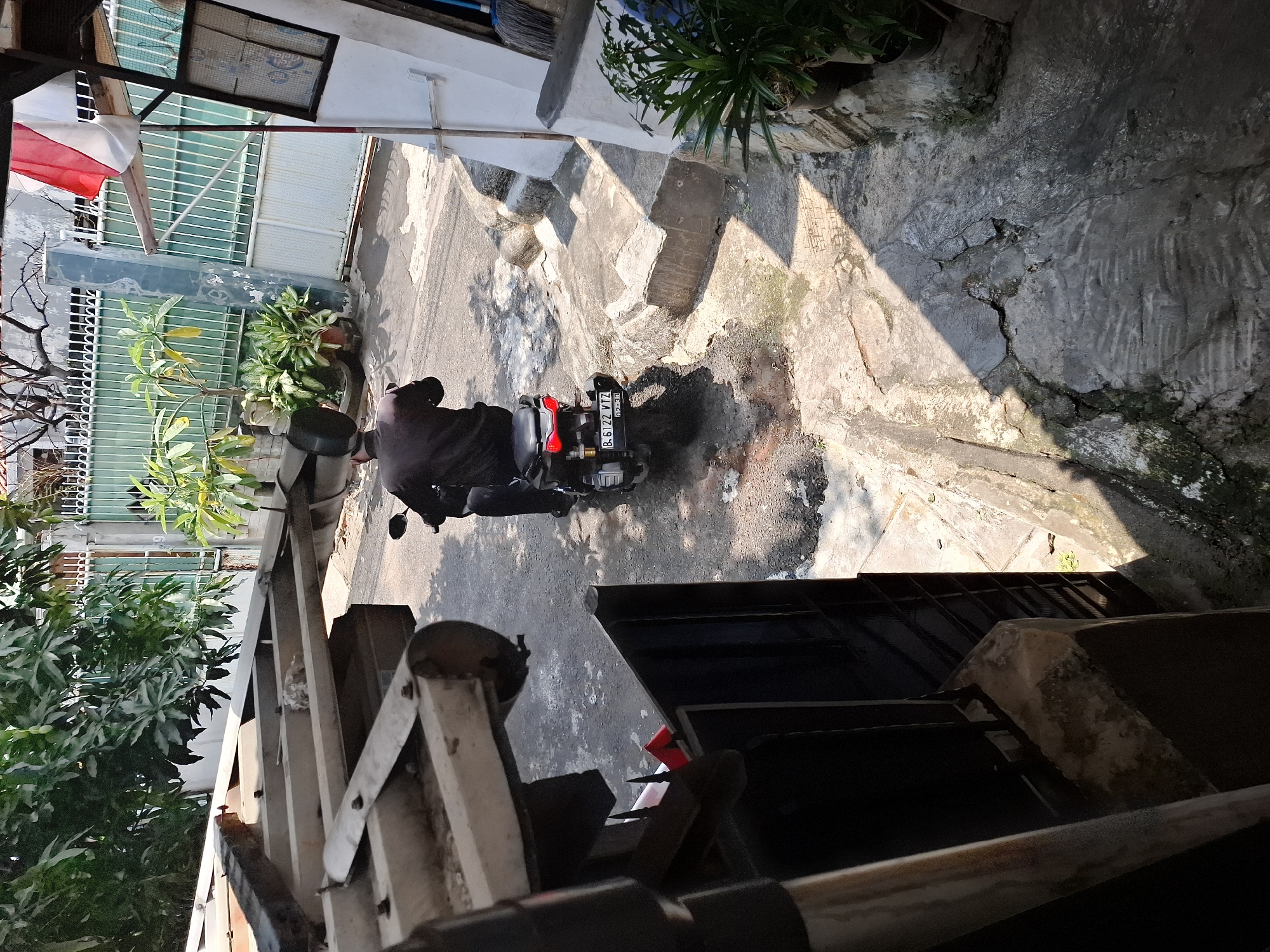 A person wearing a black long-sleeved shirt and black pants is riding a black motorcycle. The motorcycle has a license plate that reads "B. 6122 VTZ". The scene appears to be a street in a residential area in West Jakarta, Indonesia, with a building to the right that has an Indonesian flag displayed. The street is paved with cracked concrete, and there are some plants and a broom visible. The image is taken from a slightly elevated perspective, looking down at the street.