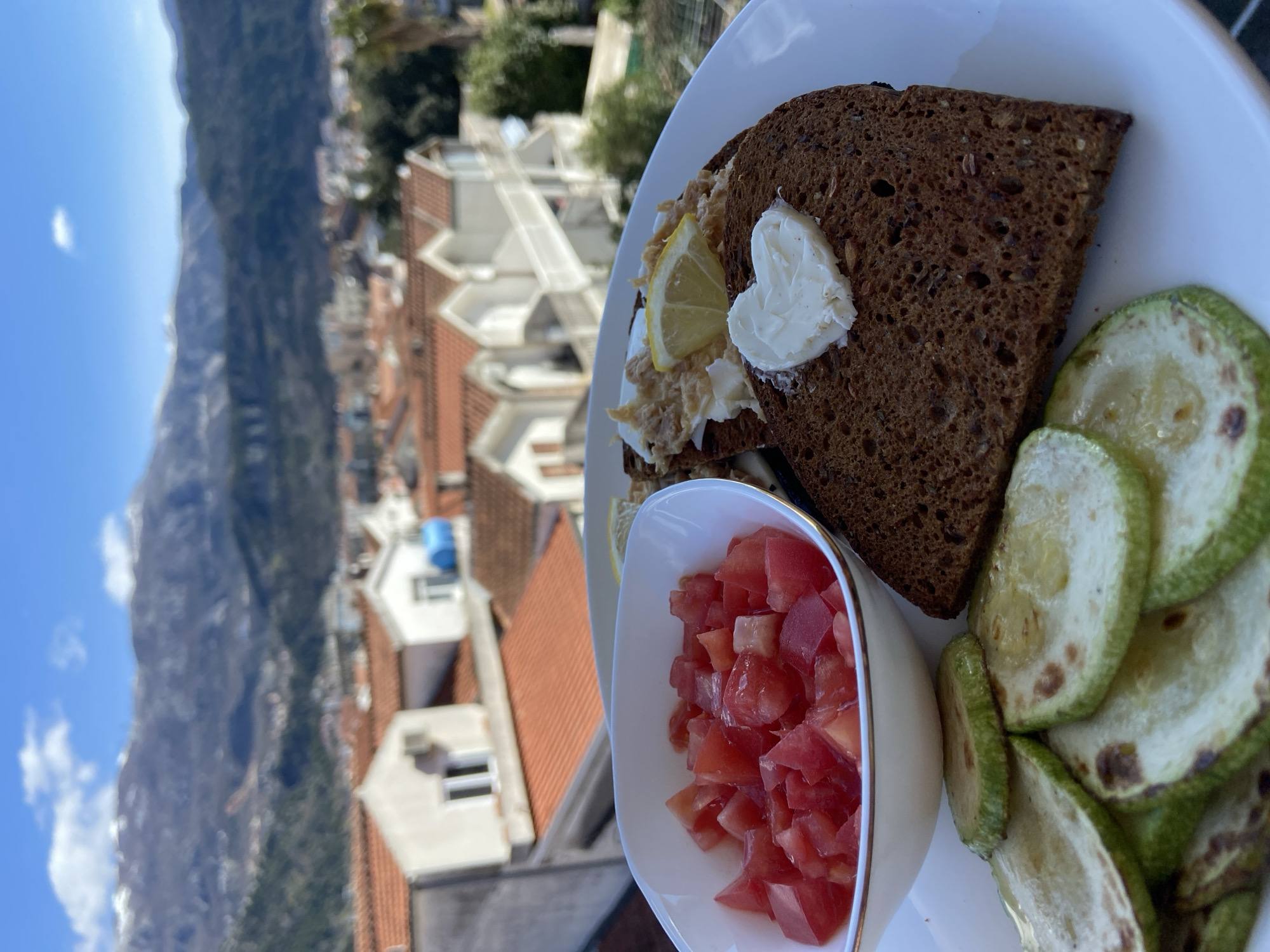 A top-down, close-up shot shows a plate of food consisting of dark bread with a heart-shaped pat of butter, tuna salad with a lemon slice, a small bowl of diced tomatoes, and sliced, grilled zucchini. The plate is on a balcony, with a view of red-roofed buildings of Budva, Montenegro, in the background. A large mountain is visible in the distance, with a blue sky above. There are no people visible in the frame.
