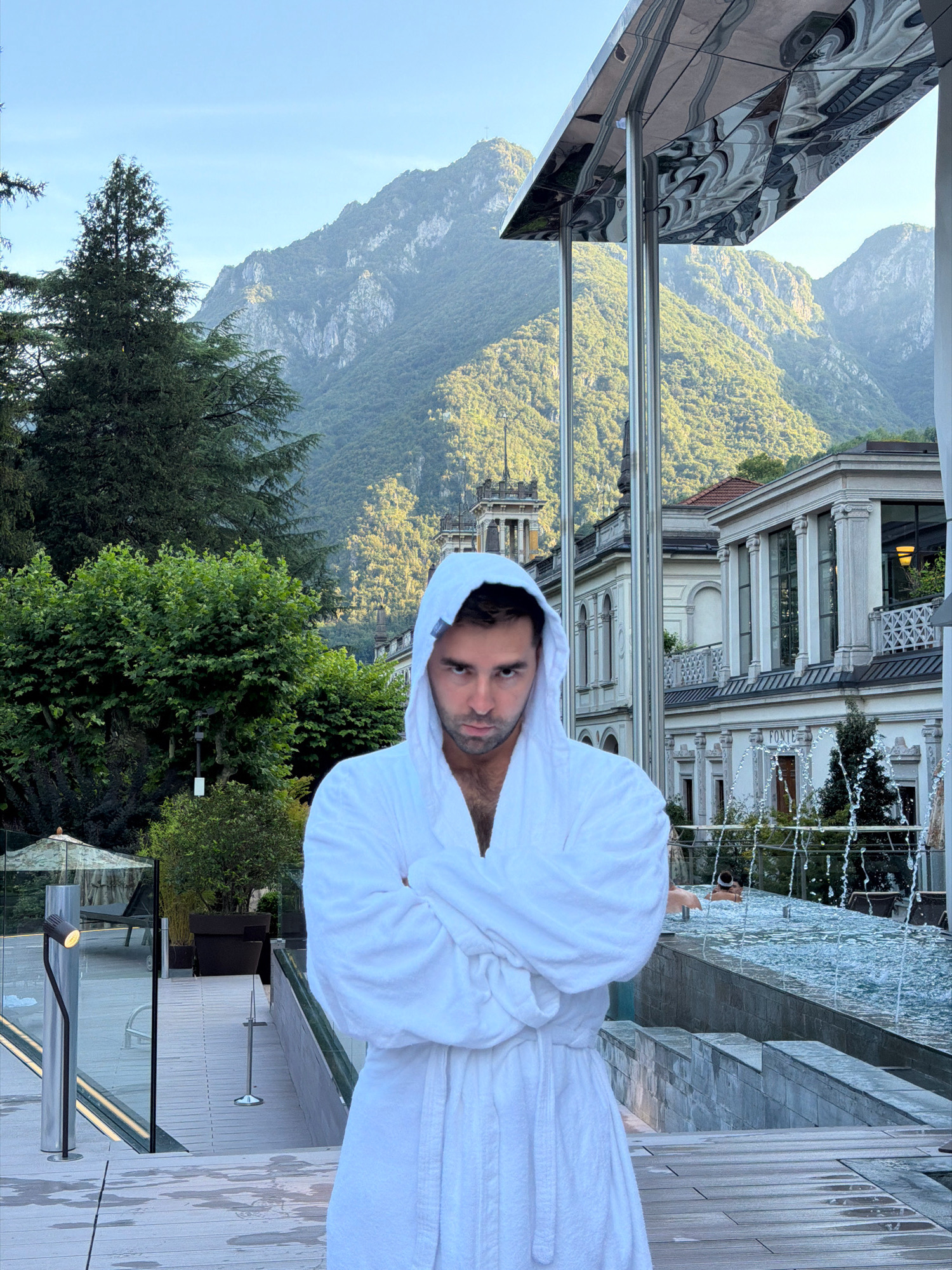 A man wearing a white bathrobe with the hood up is standing in the foreground, facing the camera with his arms crossed. He has dark hair and a serious expression. The background features a luxurious outdoor spa area with a pool and fountains. Beyond the spa, a grand building with classical architecture and a large, ornate tower is visible. In the far background, lush green mountains rise under a clear blue sky. The scene appears to be set in San Pellegrino Terme, Italy, known for its thermal baths and historic spa resort. The lighting suggests it is daytime, possibly late afternoon or early evening.