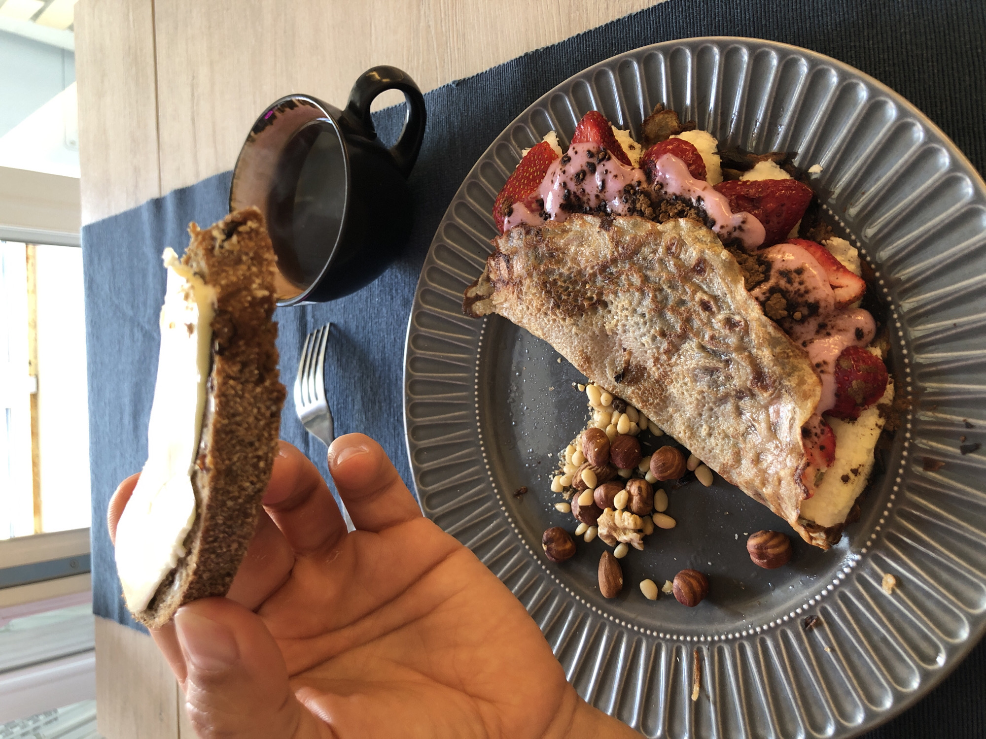 A hand is holding a slice of dark bread with white cheese on it, hovering over a cup of black coffee. In the foreground, a gray plate holds a folded crepe filled with strawberries, banana, and a pink creamy sauce, sprinkled with chocolate shavings and nuts. The setting appears to be a table with a blue placemat, likely indoors, with natural light coming from a window. The coffee is steaming.