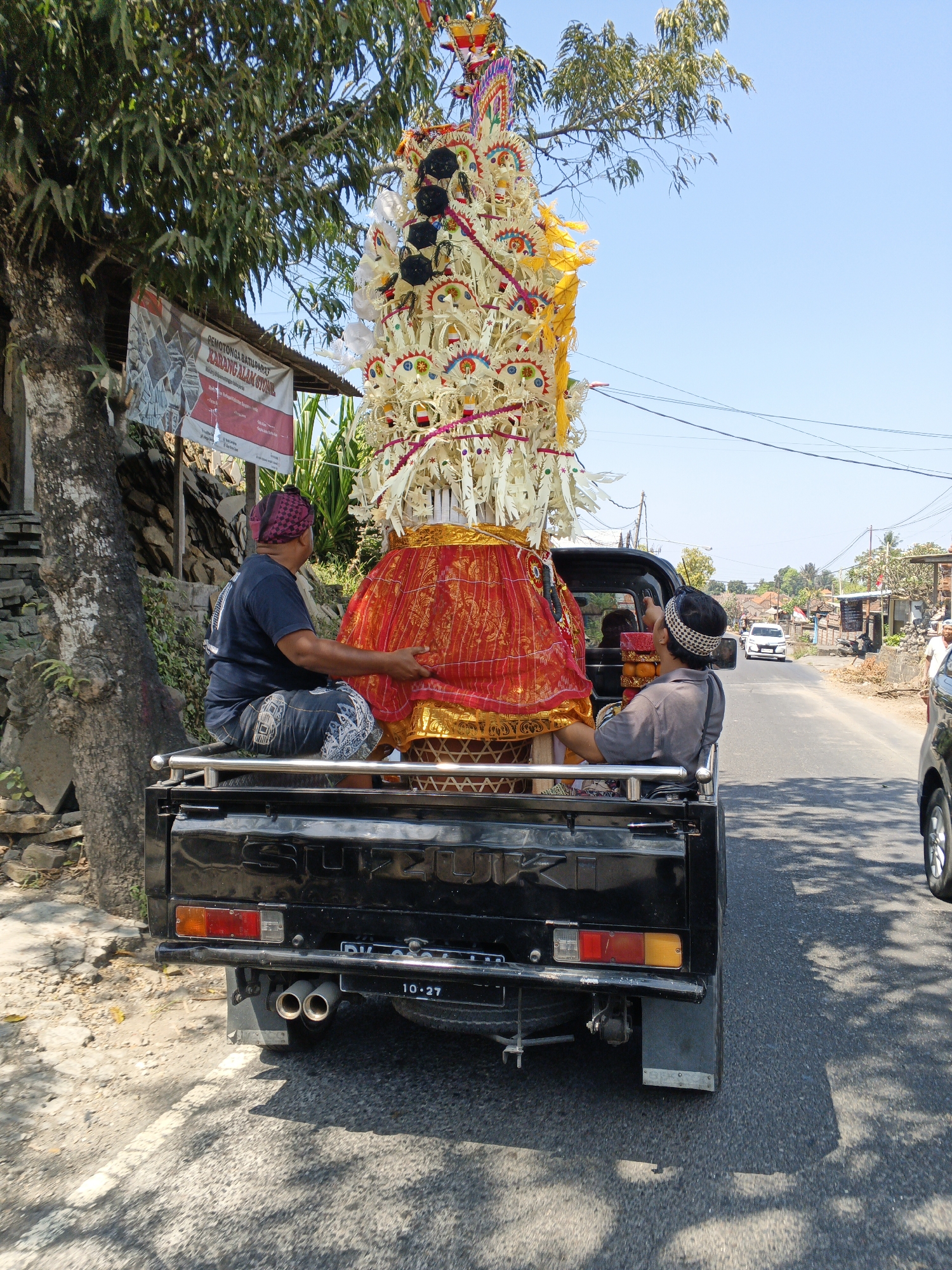 In Sukawati, Indonesia, two men are seated in the back of a black Suzuki pickup truck, transporting a large, ornate, multi-tiered structure. The structure is elaborately decorated with white and yellow elements, topped with black circular accents, and draped with a vibrant red and gold fabric skirt. The truck is driving on a paved road, with trees and buildings lining the sides. The sky is clear and blue, suggesting a sunny day. One man, wearing a dark t-shirt and patterned shorts, is leaning forward and appears to be adjusting or holding the red fabric. The other man, wearing a grey shirt and a patterned head covering, is looking upwards, possibly at the structure or the sky. In the background, other vehicles and houses can be seen, indicating a populated area. The overall scene depicts a procession or transportation of a significant cultural or religious object within an Indonesian town.