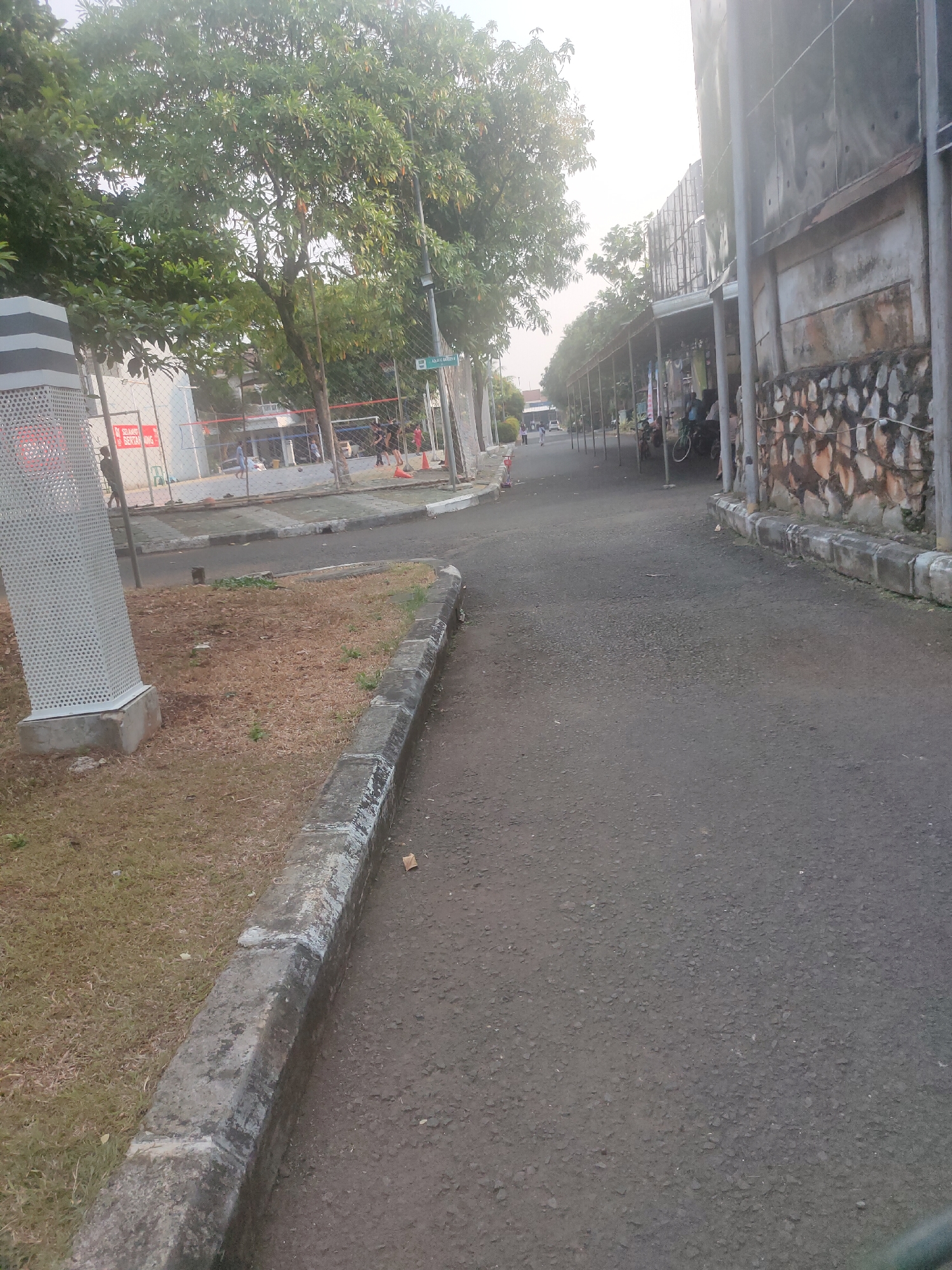 The image depicts an outdoor scene on a paved pathway next to a grassy area with a concrete curb. On the left, a tall, perforated rectangular structure is visible, along with a chain-link fence surrounding a sports court where several individuals are engaged in activities. Further down the pathway, a street sign with "AD. ADH Garden N" is mounted on a pole. To the right of the pathway, a building with a textured stone wall and a covered outdoor area supported by poles is present. In this covered area, several people and parked bicycles can be observed. The overall environment suggests a public space with recreational facilities and a marketplace or vendor area. The lighting indicates it may be late afternoon or early morning, with the sun casting a soft glow. The location is identified as Lambangjaya, Indonesia.