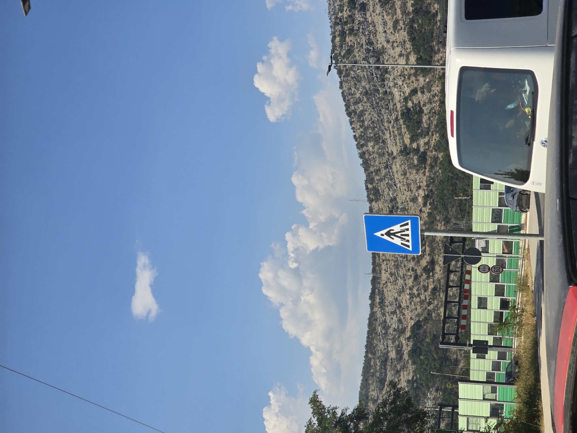 A bright, sunny day in Podgorica, Montenegro, is captured in this image. The sky is a vibrant blue, dotted with fluffy white clouds. In the foreground, the hood of a red car is visible, suggesting the perspective of someone inside a vehicle. To the right, the rear of a white van is prominently featured, with its windows reflecting the surrounding environment.
The middle ground of the image presents a pedestrian crossing sign, a common sight in urban areas. Behind the sign, a long, green barrier stretches across the landscape, punctuated by rectangular windows. This barrier likely serves as a sound or visual buffer, possibly along a road or railway. Two circular signs are affixed to a pole near the pedestrian crossing sign; one reads "4.5m" and the other "60," indicating height and speed restrictions respectively. Further in the distance, a hilly, rocky landscape covered with sparse vegetation rises, with a few power line towers visible on its slopes. A street lamp stands tall on the right side of the frame, its dark silhouette contrasting against the bright sky.
The overall impression is one of a typical roadside scene in a city, with elements of traffic, infrastructure, and the natural landscape coming together under a clear sky.