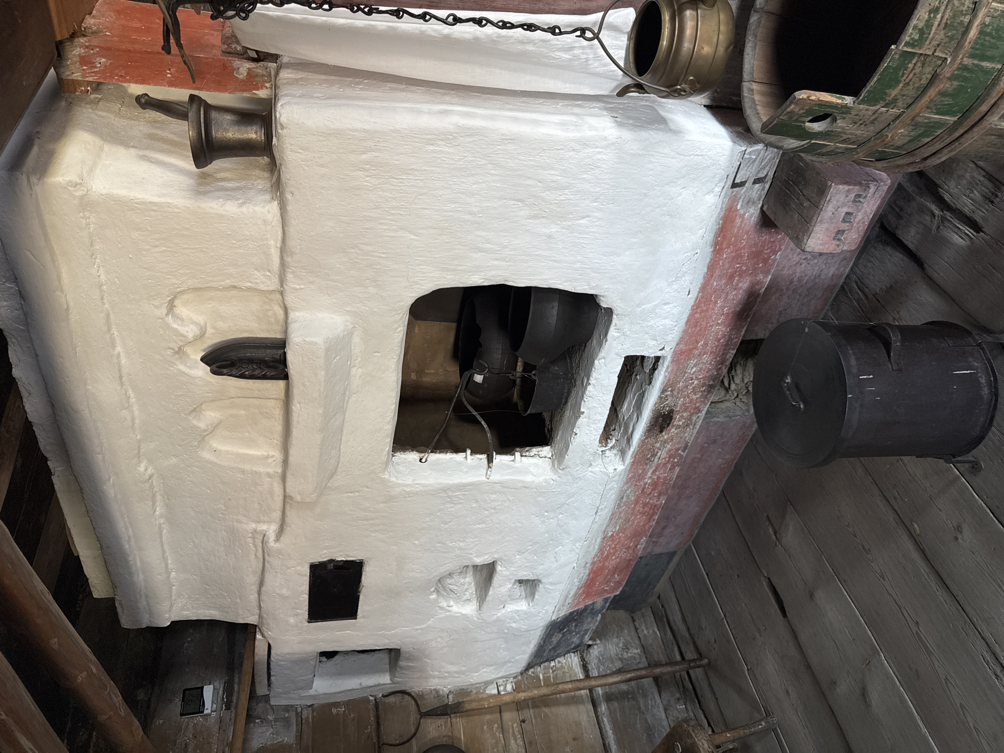 In the village of Boyarshchina, Russia, a rustic, whitewashed stove dominates the scene within a wooden structure. The stove, textured and uneven, features multiple openings and decorative niches. A small, dark metal door is set into its upper left side, and a larger rectangular opening below it reveals several dark cast-iron pots, suggesting recent use.
The stove's base is painted with a red band, overlaid with black and grey sections, showing signs of age and wear. A weathered wooden floor, made of wide planks, extends across the foreground. To the left, a long-handled metal tool with a fork-like end rests against the stove. On the stove's upper shelf, a metal mortar and pestle sit next to a decorative cast-iron object that resembles an old-fashioned iron.
To the right, a white cloth hangs near the stove, partially obscuring a portion of the structure. A brass pot, suspended by a chain, hangs from above. Further to the right, a large, green-banded wooden bucket is visible. In the foreground, a black metal cylindrical container with a lid stands on the wooden floor. Above, exposed wooden beams form the ceiling of the room. A digital thermometer is mounted on a wooden wall to the left.
