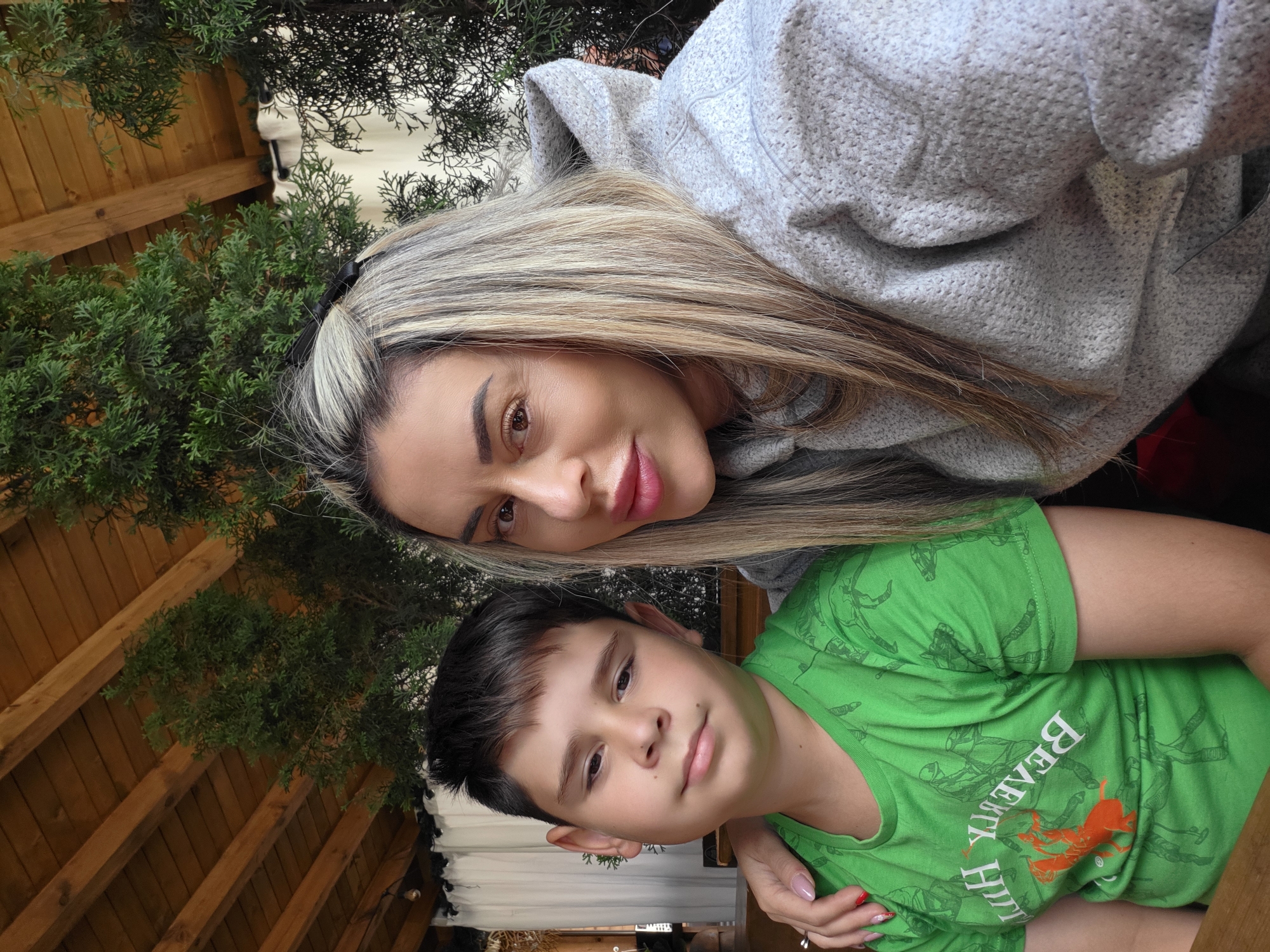 A selfie shows a woman and a young boy posing for a photo. The woman, on the right, has blonde hair and is wearing a gray hoodie. The boy, on the left, has short, dark hair and is wearing a green t-shirt with an orange horse graphic and white text. The woman's arm is around the boy's shoulders. They are seated outdoors under a wooden roof structure, with green foliage visible in the background. The scene suggests a casual outdoor setting, potentially a restaurant or cafe.