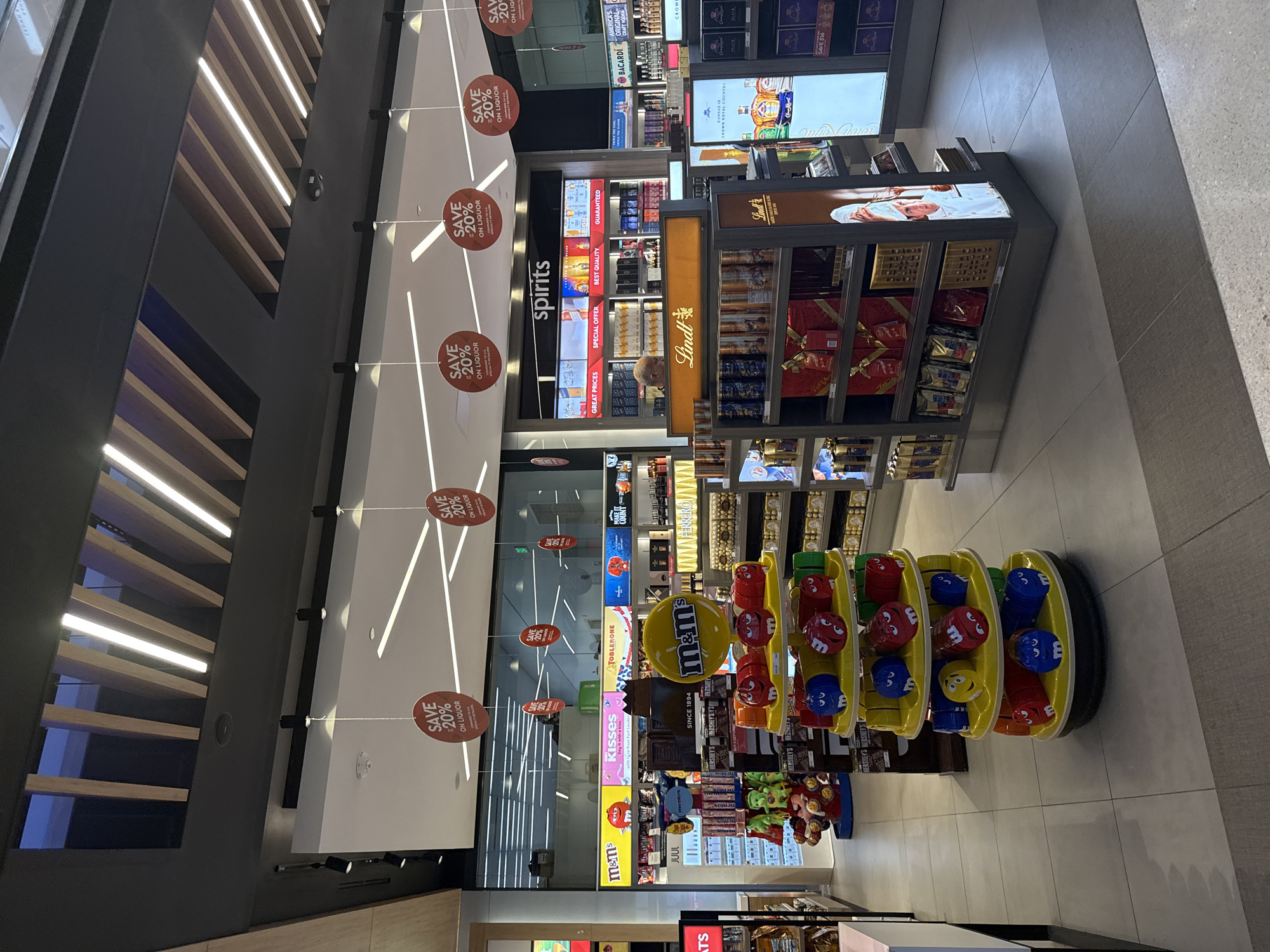 The image shows a retail store, likely in an airport, with a focus on liquor and candy. The store is well-lit with a modern design featuring a ceiling with alternating light and dark wood panels. Prominent signage advertises "SAVE 20% ON LIQUOR." Shelves display various liquor brands like Bacardi and Crown Royal, along with Lindt chocolates. An "m&m's" display stands out with colorful bins containing candy. The presence of Fort Lauderdale on a product suggests the location is in that city. There are no people visible in the immediate foreground.