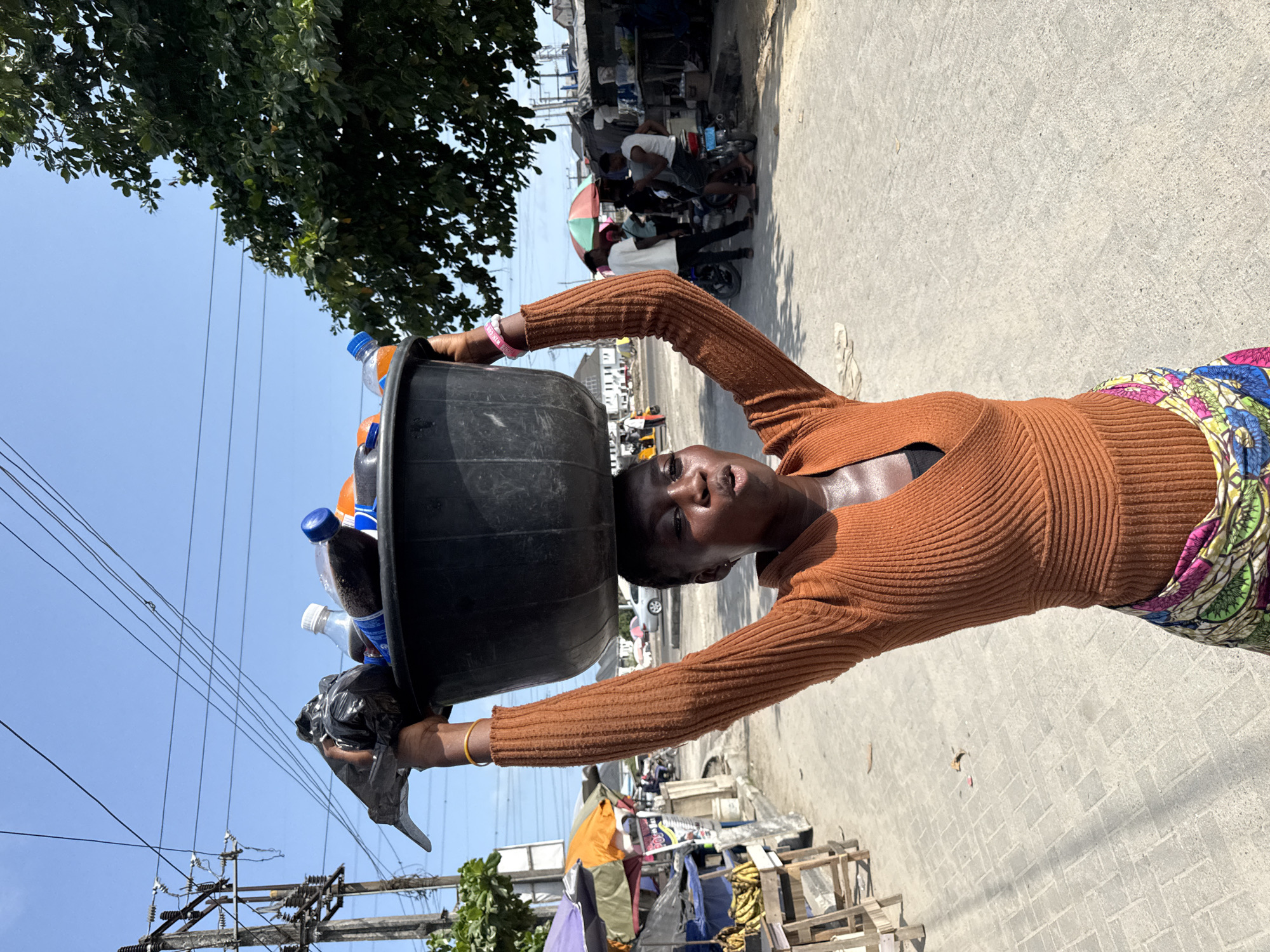 In Igboefon, Nigeria, a woman balances a large black basin filled with plastic bottles and a black plastic bag on her head. She is wearing a long-sleeved rust-colored shirt and a brightly colored skirt with floral patterns. Her arms are raised to support the basin. A gold bracelet is visible on one wrist and a pink bracelet on the other. The background shows a busy street scene with people, motorcycles, and makeshift stalls or shops. Power lines and trees are visible in the sky. The pavement is concrete. The light is bright, suggesting a sunny day.