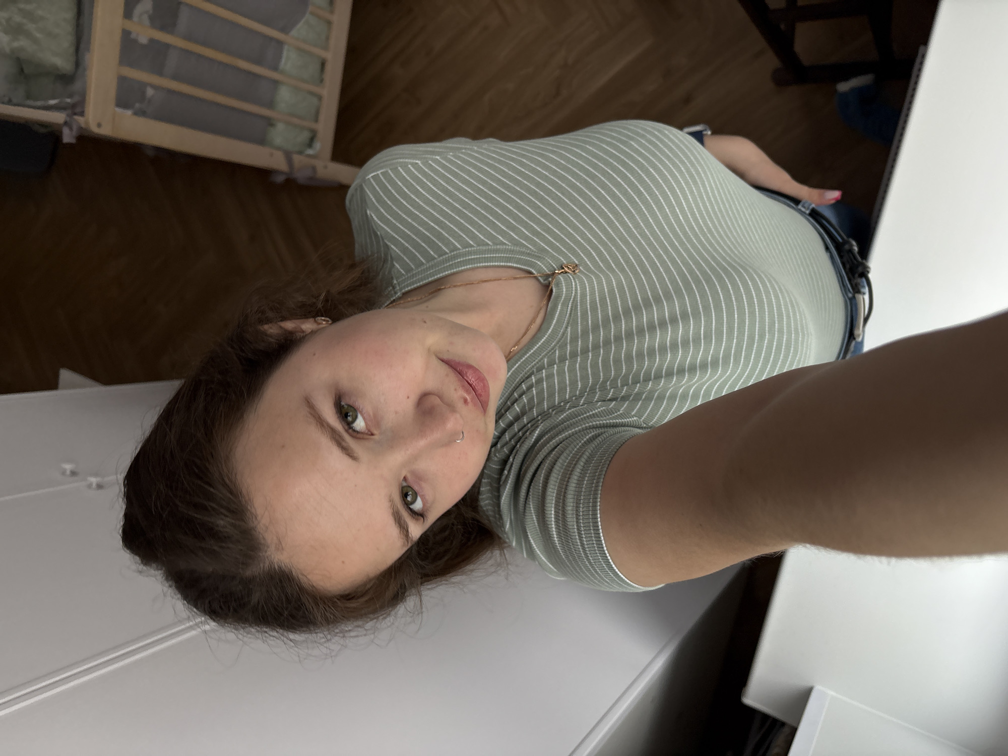 A young woman is shown in a selfie-style photograph, appearing from a high angle. She is wearing a light green and white striped t-shirt and a thin gold necklace. She has brown hair and fair skin. The background includes wooden floors, a crib with a grey interior, and white furniture. Given the request's specification of Izhevsk, Russia, one can infer the photo was taken in a residential interior in that city, although this cannot be definitively confirmed from the image alone. There are no evident actions, interactions, or visible objects associated with the location beyond what is seen.
