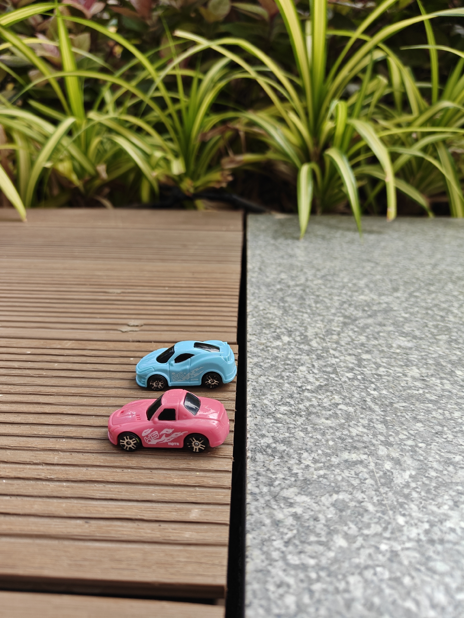 The image shows two toy cars, one blue and one pink, positioned on a wooden surface with a grey tiled surface adjacent to it. The background features greenery. The location is identifiable as Mumbai, India based on plant species typical of the area.