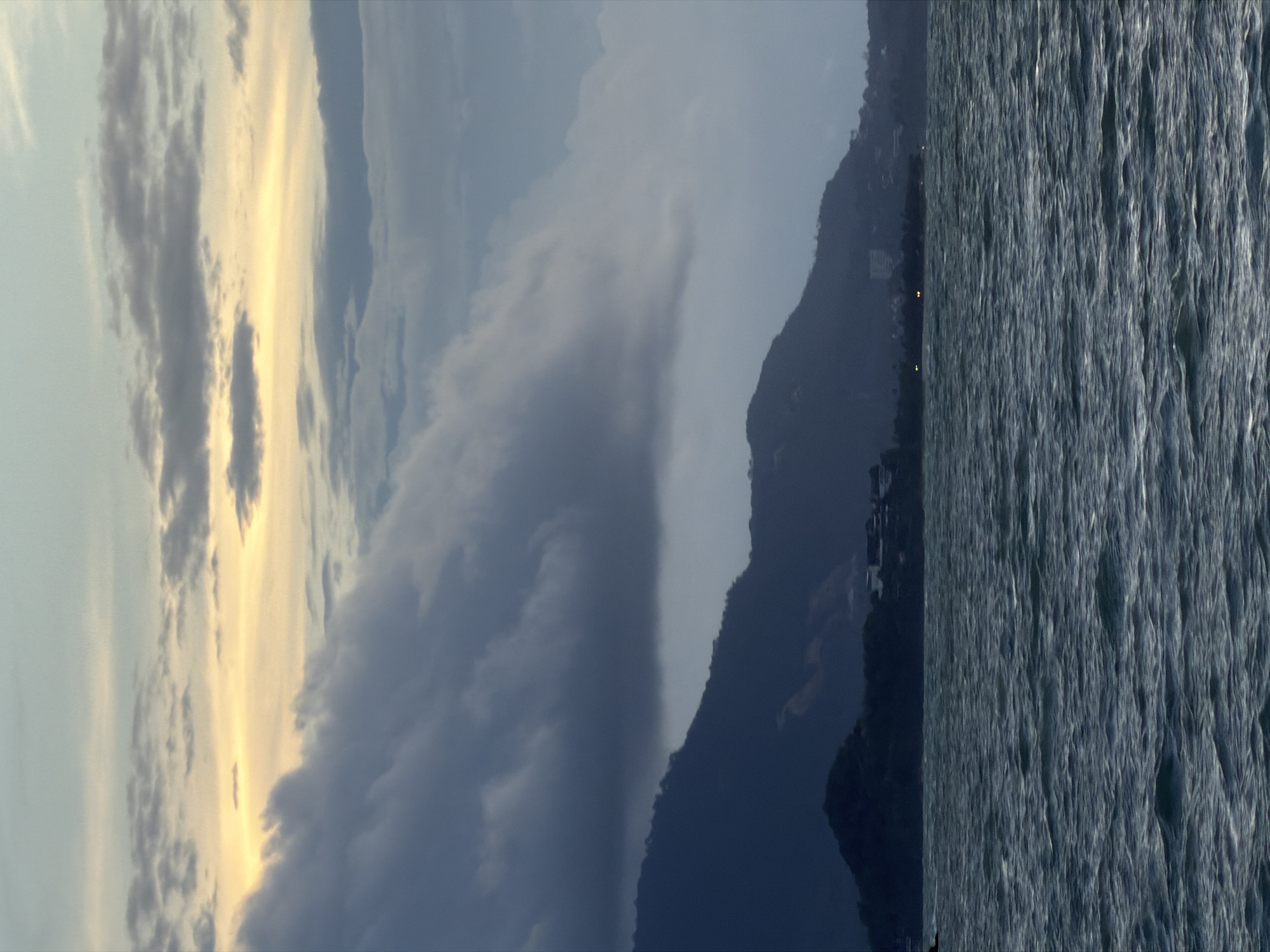 The image depicts a scenic coastal view in Ko Samui District, Thailand, likely during the late afternoon or early evening. The foreground is dominated by the dark, choppy waters of the sea, with visible waves creating a textured surface. In the middle ground, a silhouetted mountain range rises from the water's edge, its dark contours contrasting with the lighter sky. Along the base of the mountains, a low-lying area with scattered lights suggests human settlement, with buildings and possibly trees visible. The background features a dramatic sky filled with various cloud formations. Dark, heavy clouds hang low over the mountains, while higher up, lighter, wispy clouds are tinged with a warm, golden light, indicative of sunset or sunrise. The overall atmosphere is serene and somewhat moody, highlighting the natural beauty of the island. No people or specific actions are discernible in the image.