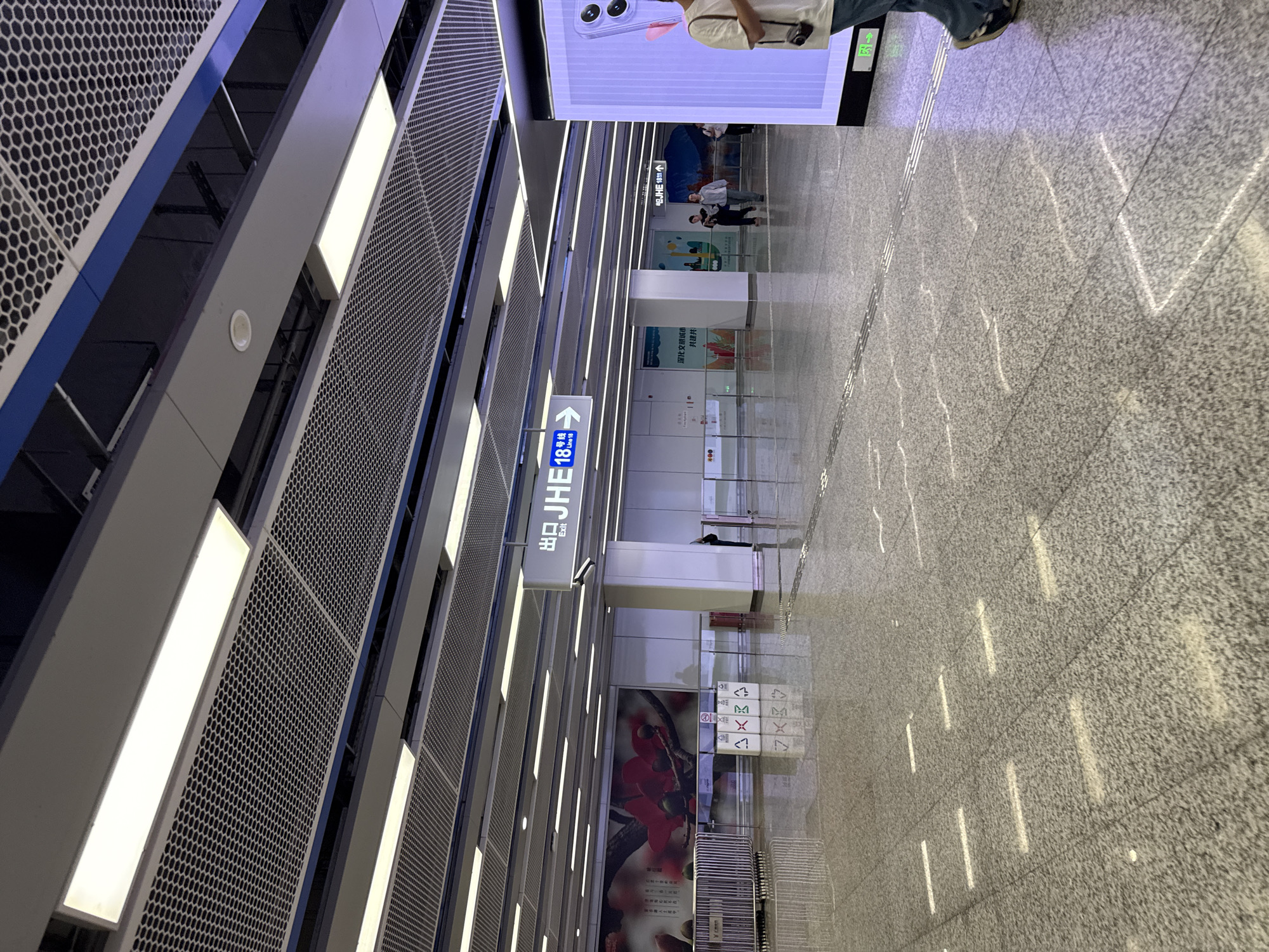 The image shows an interior view of a modern subway station in Haizhu District, China. The ceiling features a series of linear lights and a perforated metal panel system. The floor is a polished, speckled granite tile.
In the background, several signs are visible. One prominent sign, with white lettering on a blue background, indicates "出口 JHE Exit" and "18号线 Line 18" with an arrow pointing to the right. Further down the platform, another sign in Chinese and English points towards "JHE 18号线 Line 18." There are also large advertising displays or murals on the walls.
Several people are present, appearing to walk through the station. One individual is prominently featured in the foreground on the right, partially obscured by a large digital display. Other individuals are seen walking in the middle ground.
The station appears clean and well-maintained, with reflections on the polished floor indicating a bright, indoor environment. Various amenities like trash receptacles and safety barriers are also discernible. The overall impression is of a functional and contemporary public transportation hub.