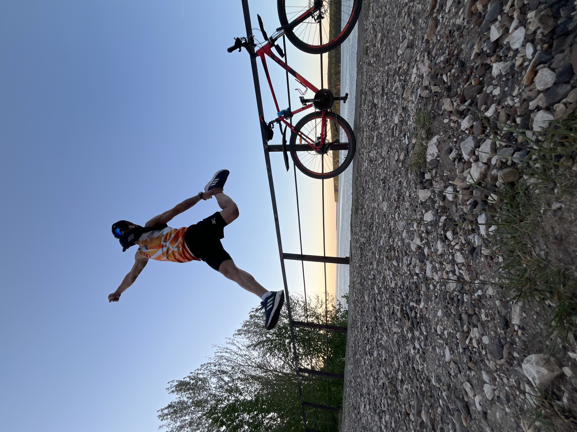 The image shows a man in an orange and white athletic top, black shorts, and black and blue athletic shoes, striking a yoga-like pose outdoors. He is holding his right foot with his right hand, extending his left arm outward, and wearing a cap and glasses. Behind him, a red bicycle is propped against a metal railing. The railing borders a rough, rocky surface, possibly a path or embankment. Beyond the railing and the rocks, there is a body of water, suggesting a river or lake, with a hint of sunset or sunrise on the horizon. Green trees are visible below the railing. The image is angled as if the camera was tilted sideways, and the location is likely in Chaikovsky, Russia, given the prompt.