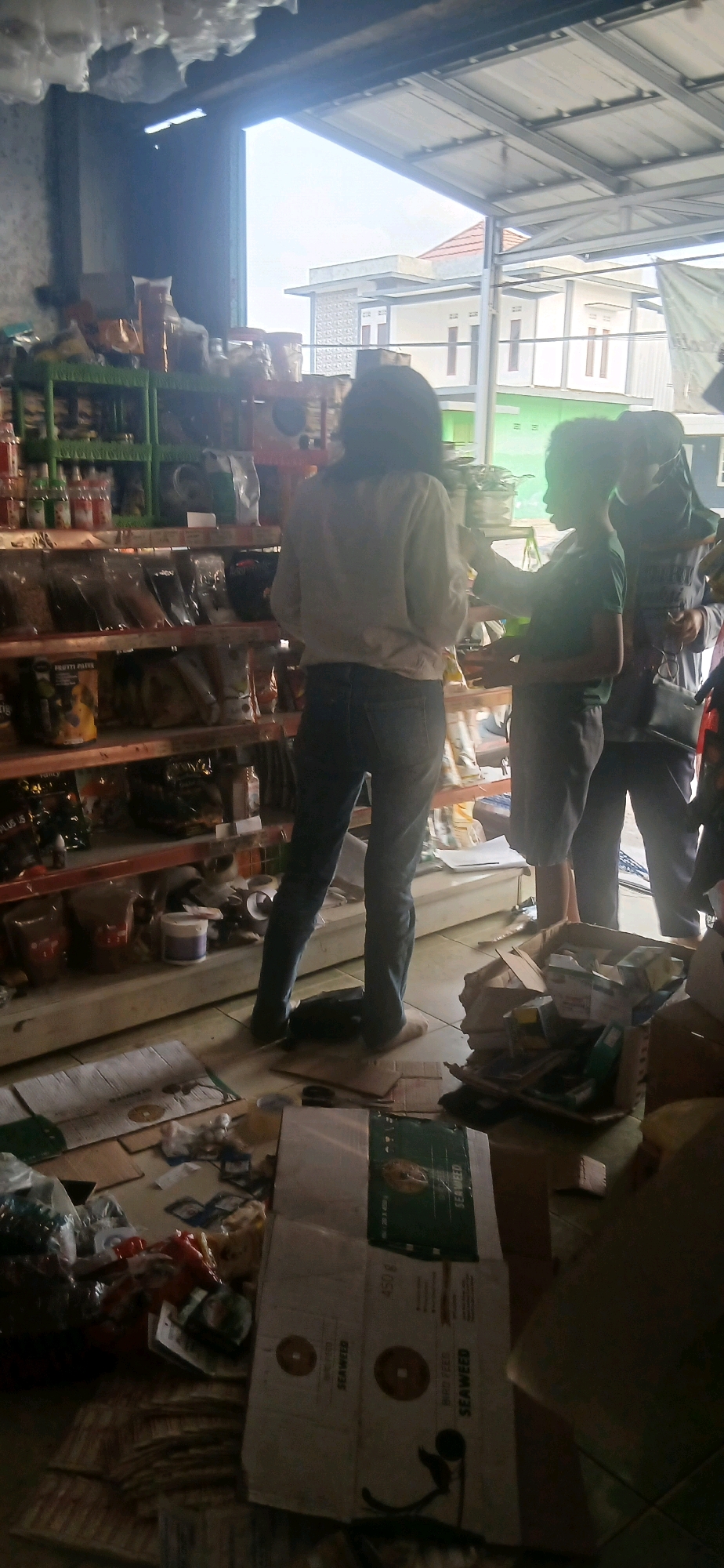 In Banar Joyo, Indonesia, a dimly lit retail space is observed with shelves stocked with various products, possibly food items and household goods. The floor is cluttered with cardboard boxes, packaging, and scattered items, suggesting a disorganized or recently stocked environment. In the foreground, the legs of a person wearing blue jeans and light-colored footwear are visible, standing on the cluttered floor. To their right, another person, also wearing jeans, is partially visible, reaching towards an object on a shelf. Further to the right, a third individual, wearing a hijab, is seen with their back to the camera, holding a dark bag. The background reveals an open-air structure with a metal roof, looking out onto buildings with white facades and a light blue sky.