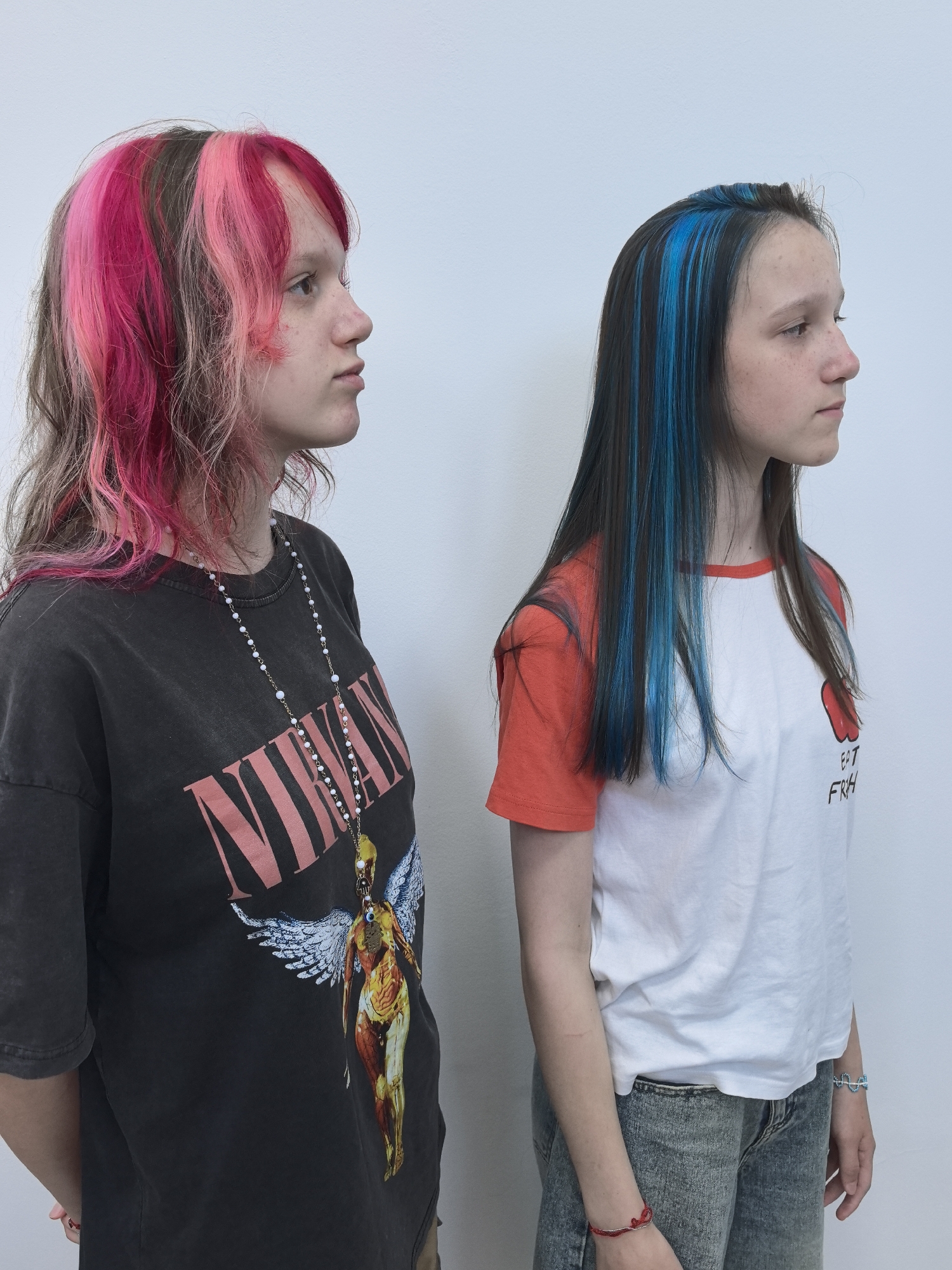 Two young females stand side-by-side in front of a white wall. The female on the left has multi-colored hair, with streaks of pink and brown. She wears a dark t-shirt with a graphic print, and a long necklace with white beads. The female on the right has dark hair with vibrant blue streaks. She wears a white t-shirt with an orange collar and sleeves, and denim jeans. The location is identified as Chaikovsky, Russia.