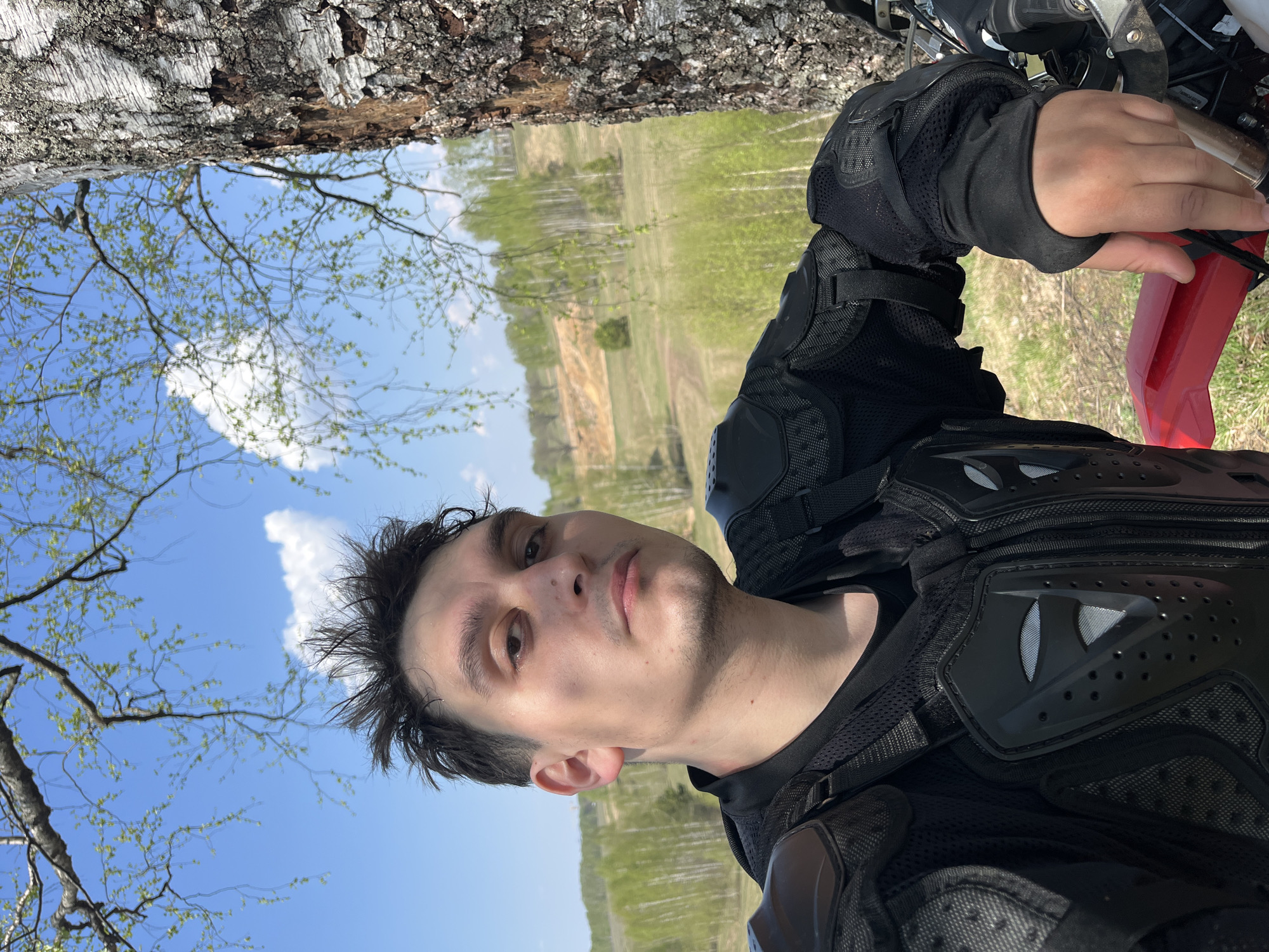 A man is pictured in a selfie shot outdoors, with a background containing a field or forest and a bright blue sky with sparse clouds, possibly in the area of Новочувашский, Russia. The man is wearing a black protective motorcycle suit, including shoulder pads. He appears to be on a motorcycle. A tree trunk is visible in the background.