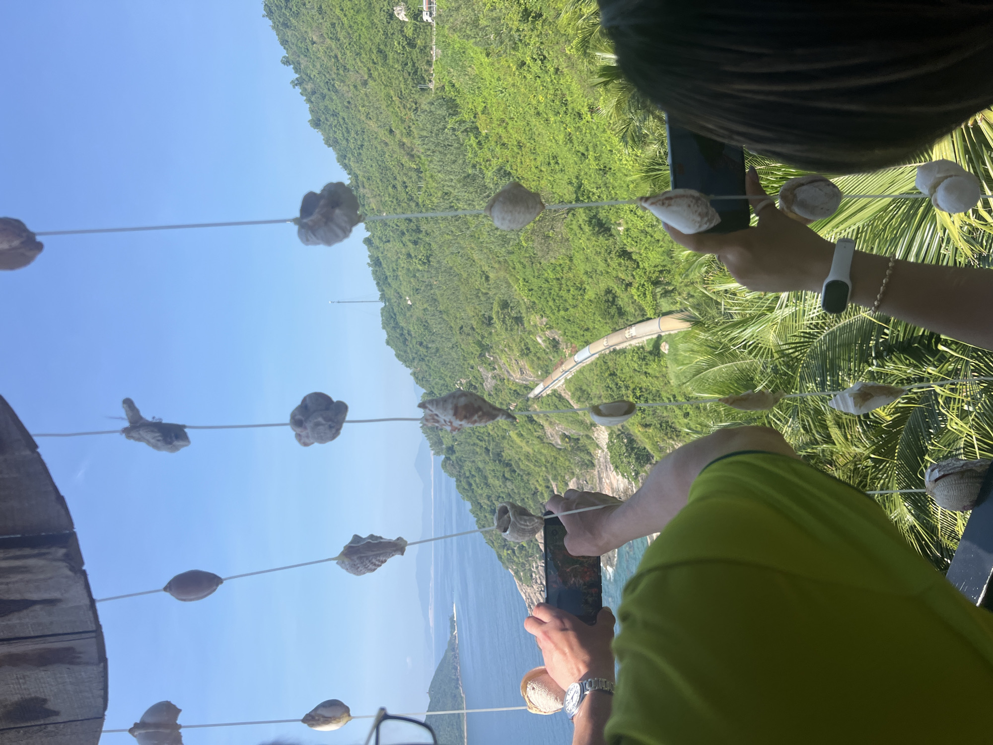 The image depicts a vibrant coastal mountainous landscape under a clear blue sky, viewed from a high vantage point in the region of Hòa Xuân, Vietnam. In the immediate foreground, a partial wooden structure suggests an overhead canopy or roof. Numerous seashell wind chimes, suspended by thin white strings, hang prominently in the foreground, creating a layered depth of field. These chimes consist of various types of light-colored seashells, including conchs and other spiral and bivalve forms.
Two individuals are partially visible, positioned in the lower foreground, each holding a smartphone horizontally as if taking a photograph or video of the expansive view. The person on the left, identified by the back of a bright lime-green t-shirt and partially visible eyeglasses, holds a black smartphone with both hands. A silver-toned wristwatch is visible on their left wrist. To their right, the second individual, with dark hair visible from the back, holds a smartphone with a blue-colored back. A white fitness tracker and a beaded bracelet are visible on their left wrist. A dark railing or ledge is partially visible beneath their hands.
Beyond the immediate foreground, a verdant mountain slope dominates the midground, densely covered with various shades of green foliage, including palm trees in the lower section. A winding light-colored road is visible on the hillside, with a section of what appears to be a protective barrier running alongside it. A white truck is partially discernible on this road in the upper right quadrant. Further down the slope on the left, a long, light-colored, tubular structure, possibly a water slide or aqueduct, curves downwards towards the coastline.
The deep blue sea occupies the left and central midground, meeting a rocky shore at the base of the mountains. Distant islands or landmasses are visible across the water, receding towards the horizon. A tall, slender communication tower stands on a distant hilltop on the left. The sky is uniformly clear and light blue, indicating fair weather conditions.