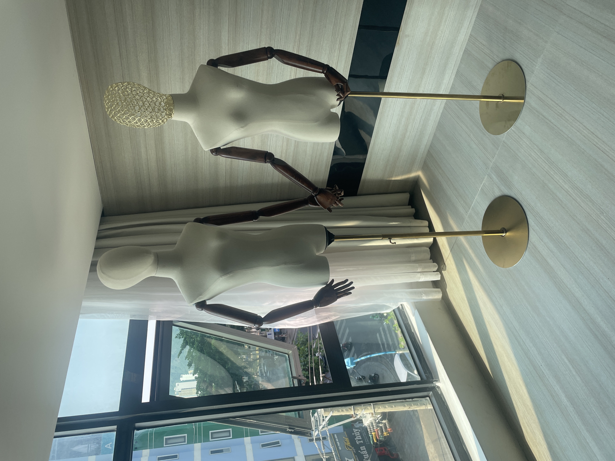 Two mannequins stand in a room with a large window overlooking a cityscape. The city of Bắc Nha Trang, Vietnam is visible through the window. One mannequin has a white body and a gold wire head, while the other is entirely white. Both have articulated wooden arms and stand on gold-colored poles with circular bases. The room has a light-colored wood-grain wall and tile floor. A curtain hangs near the lower mannequin.