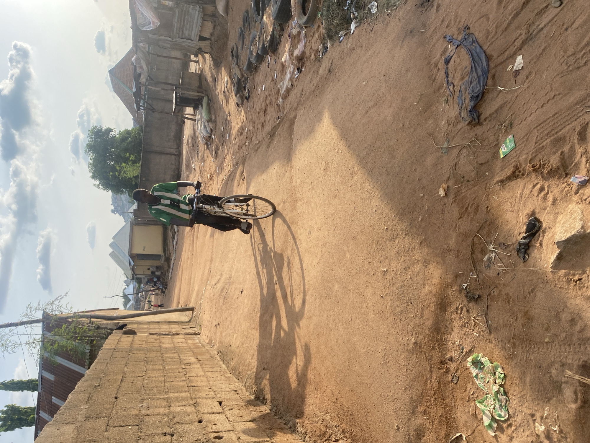 A young male wearing a green and white striped shirt and dark pants rides a bicycle down a dirt road lined with brick walls and simple structures. The sun is setting, casting long shadows on the dusty ground. Debris, including plastic bags and discarded tires, is scattered along the roadside. The environment appears to be a rural or impoverished urban area in Unknown, Unknown.