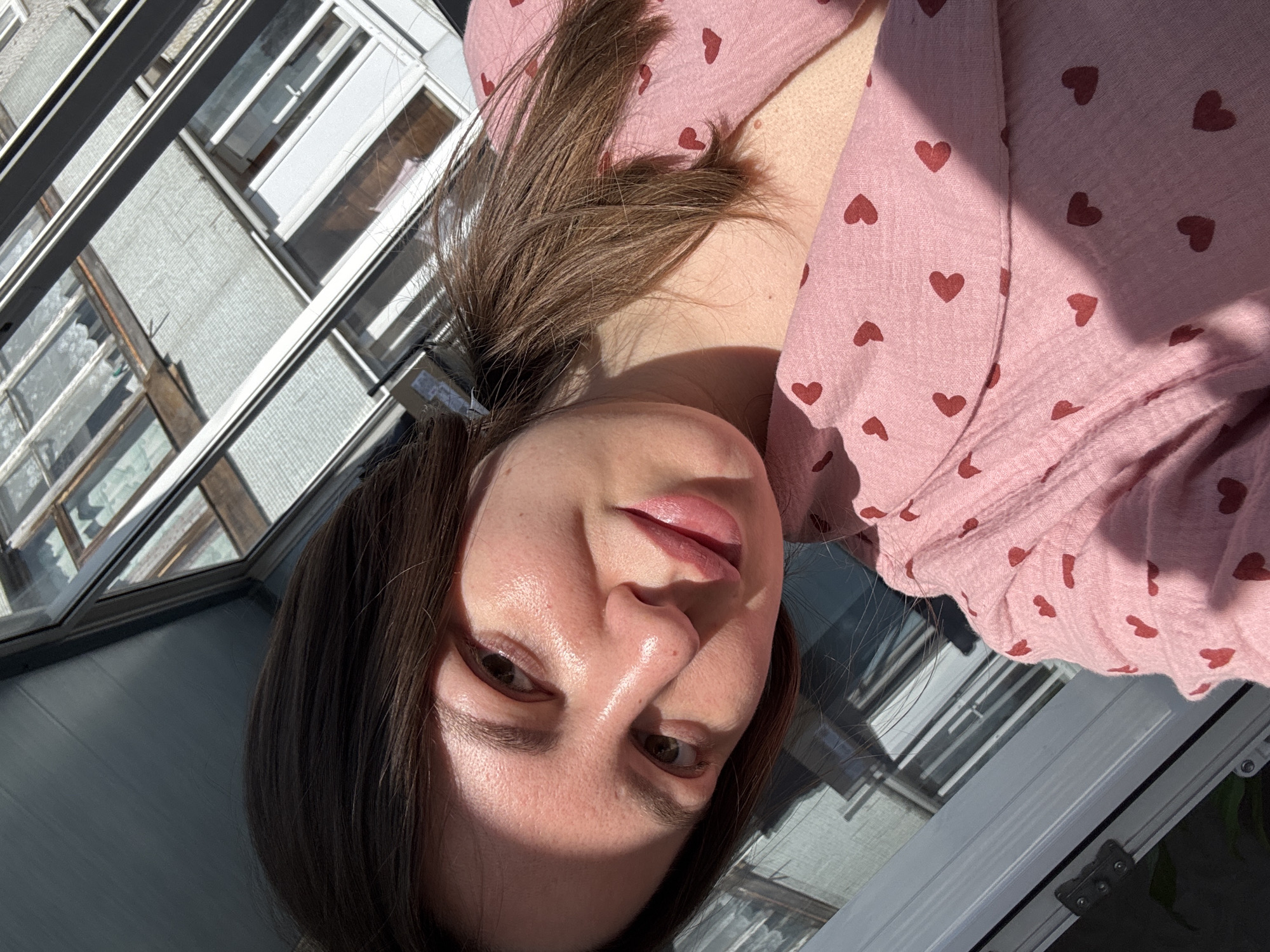 A young woman with light skin and brown hair is depicted in a close-up shot. She is wearing a pink top patterned with small, scattered red hearts. The background shows architectural details of a building, including windows with multiple panes and frames. Reflections on the window suggest an urban setting. While specific details of Orekhovo-Zuyevo, Russia are not identifiable within the image, the architectural style and the presence of what appears to be a balcony could suggest an urban residential environment. The lighting is bright, implying that the photograph was taken outdoors or near a window during daylight hours.