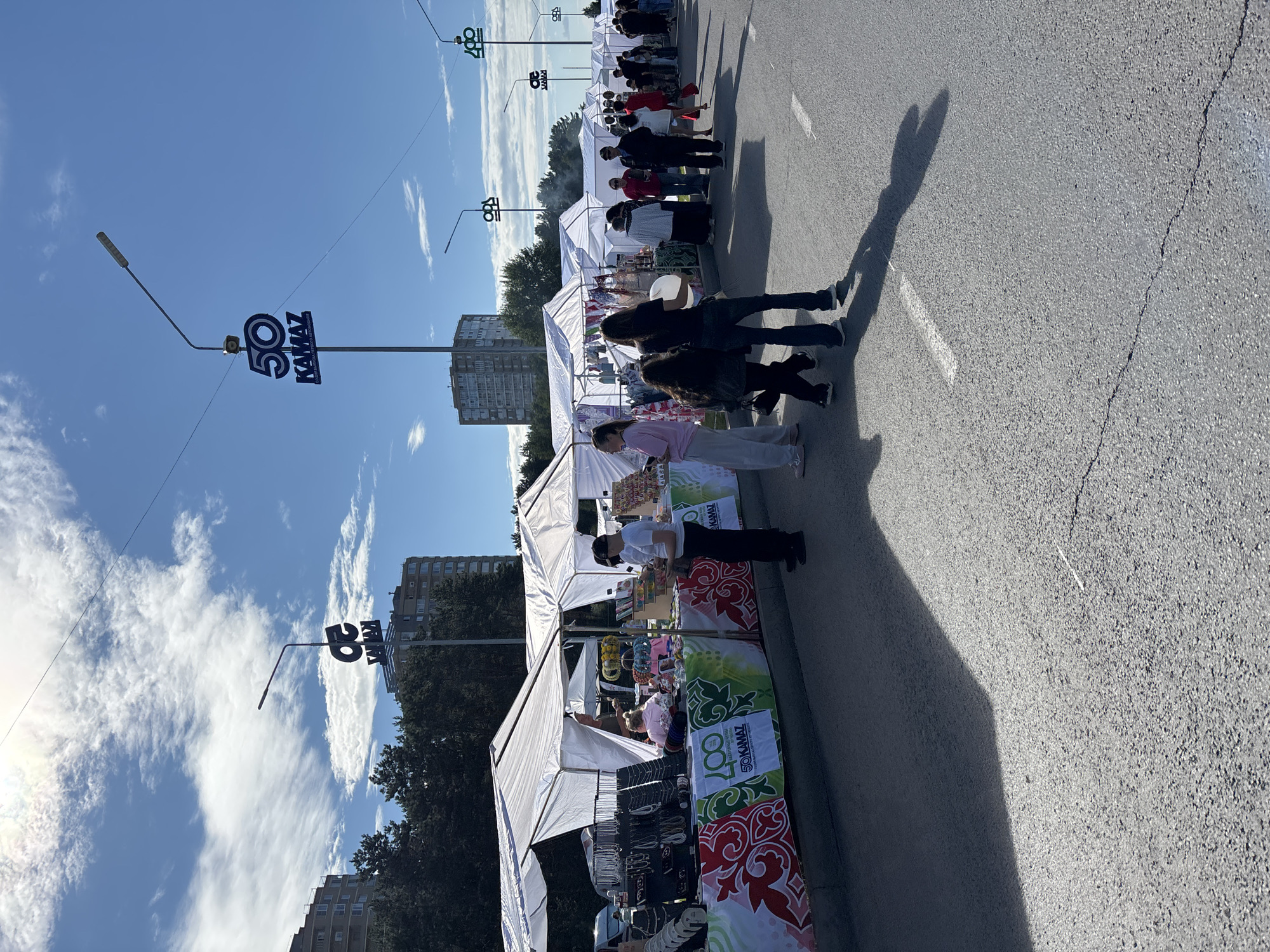 The image captures a street market scene likely taking place in Naberezhnye Chelny, Russia, indicated by the prominent "50 KAMAZ" signage. The event appears to be a celebration of KAMAZ's 50th anniversary, with multiple tents set up along a paved street. A variety of goods, including jewelry and trinkets, are displayed on tables within the tents. People of various ages are browsing the stalls and walking along the street. The weather is sunny with scattered clouds, and the sun casts long shadows. The background features apartment buildings and trees, consistent with an urban environment. The overall impression is of a community event or fair.