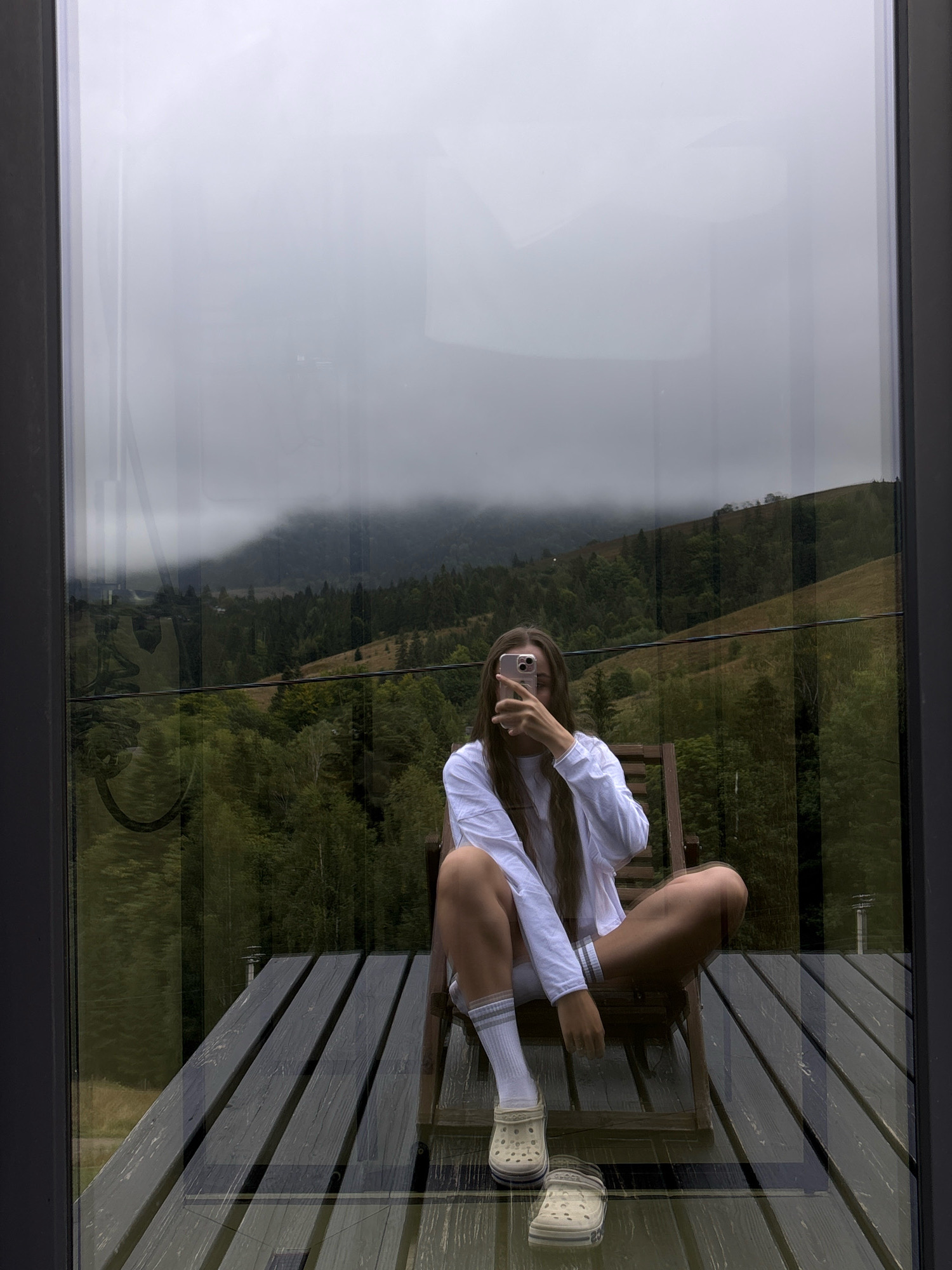 A young woman sits on a wooden chair on a deck, taking a selfie with her phone. She is wearing a white long-sleeved shirt, white shorts with striped trim, white socks, and light-colored Crocs. The deck appears to be made of dark wood planks. Behind her, through a large window, is a view of rolling hills covered in green trees and patches of grass. The sky is overcast, with clouds obscuring the mountain peaks in the distance. The reflection on the window shows a portion of the scene. This scene is located in Krasnyk, Ukraine, a village in the Ivano-Frankivsk Oblast known for its mountainous landscape.