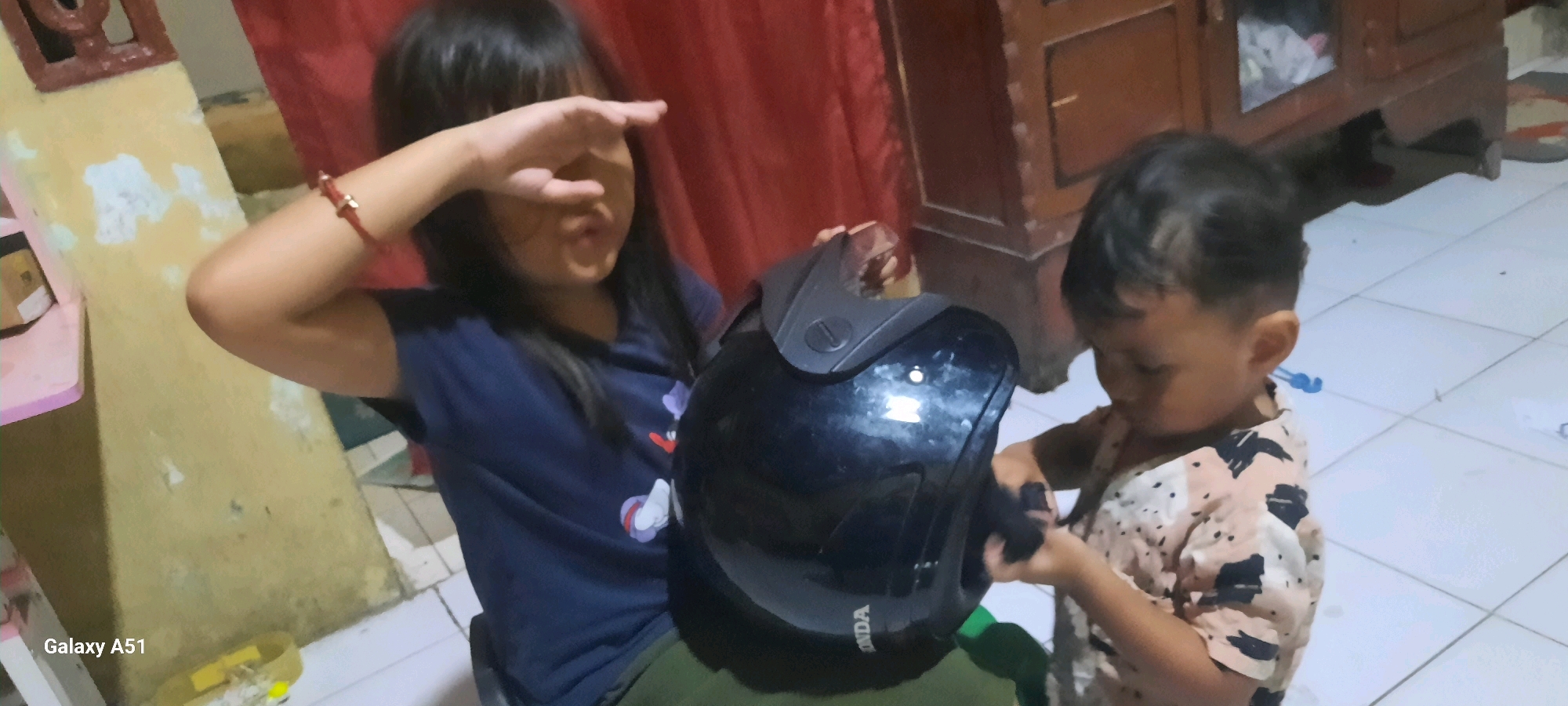 Two young children are indoors, possibly in Ploso, Indonesia. The child on the left, wearing a dark blue shirt and green pants, is covering their eyes with their left hand. They are holding a black helmet with their right hand. The child on the right, wearing a patterned shirt, is looking down at the helmet and appears to be interacting with it, holding a dark object. The background shows a red curtain, a wooden cabinet, and a tiled floor. The overall scene suggests a casual, domestic setting.