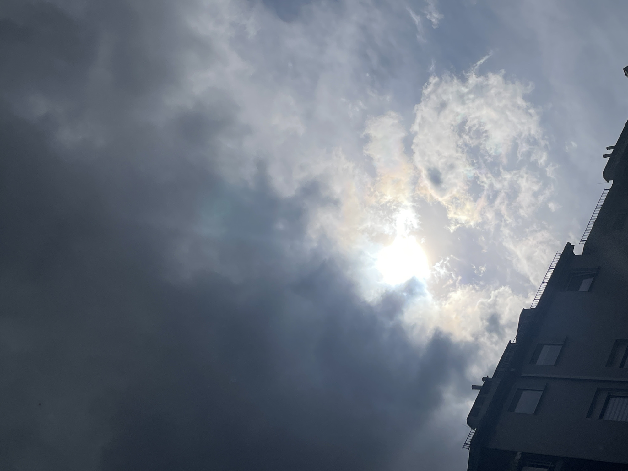 The image captures a low-angle view of a cloudy sky with the sun obscured by clouds, and a building on the right side. The clouds range from dark gray to bright white, with the sun's light filtering through, creating a radiant effect. A portion of a building occupies the lower-right corner, featuring multiple windows and architectural details. The location is Unknown, Unknown.