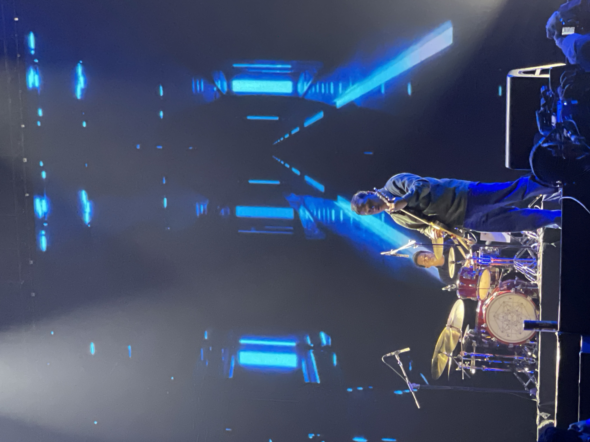 A live musical performance unfolds on a stage bathed in blue light. A guitarist, dressed in dark clothing, plays a stringed instrument. Behind him, a drummer is visible at his kit, composed of drums and cymbals, with a white design on the bass drum. A microphone stand is positioned near the drum set. The backdrop consists of geometric, blue light designs. A camera and cameraman are visible to the right of the stage, partially obscured. The location and city are indiscernible from the image.