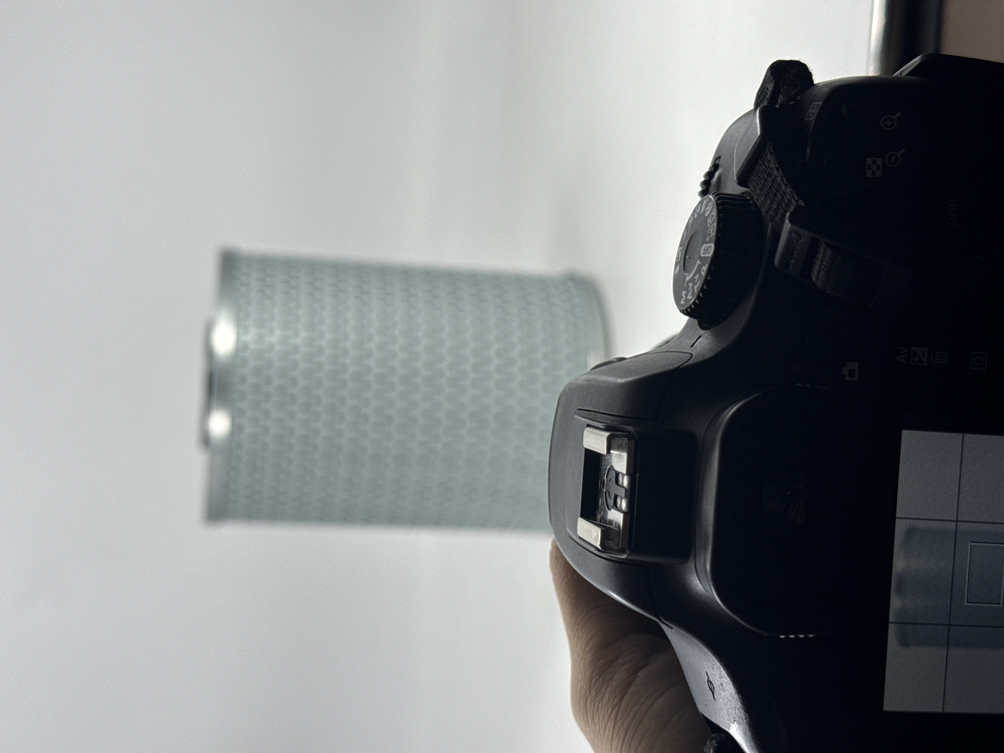A digital camera is being held to photograph a cylindrical, light-colored object with a textured surface against a white background. The camera is black and its display screen shows a grid pattern. The environment is a studio or indoor space with controlled lighting. There is no indication of a specific location like Tinaquillo, Venezuela.