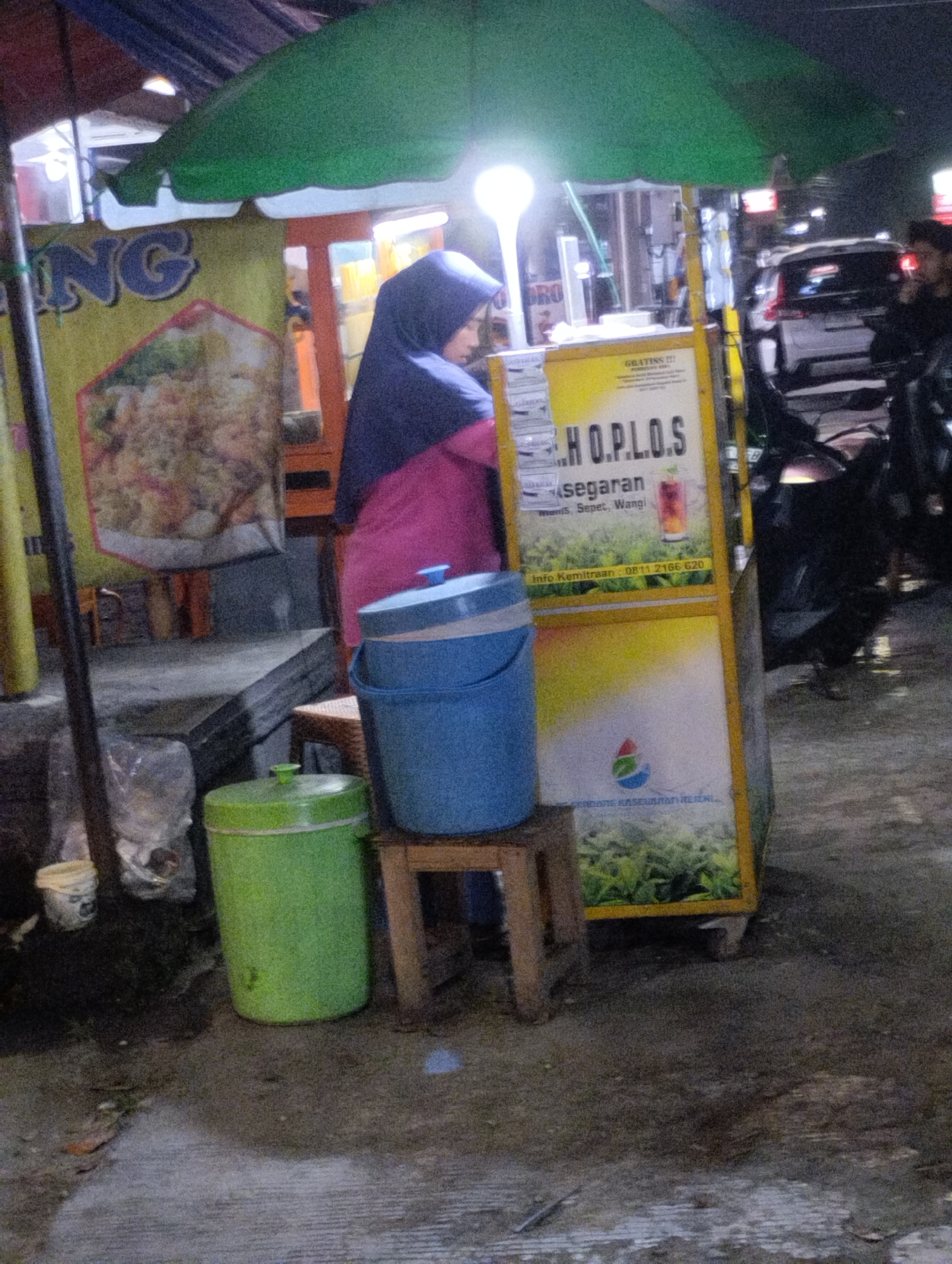 In an outdoor urban setting at night, likely in Bekasi, Indonesia, a street vendor operates a beverage stand. A woman wearing a dark hijab and a bright pink shirt stands behind the stand, tending to it. The stand itself is a yellow kiosk with various signage, one prominently displaying "H.O.P.L.O.S." in large letters and text below that reads "Segaran" which translates to "refreshing". Another sign features a logo and the text "INDONESIA RASELAA MAKI". Two large buckets, one blue and one green, are positioned next to the stand, likely for drinks or ice. The ground is paved with a rough surface, and a small white bucket sits near the green one. In the background, a white car and parts of motorcycles are visible, suggesting the presence of a road or parking area. The scene is illuminated by a bright overhead light and ambient street lighting, creating a slightly grainy, low-light effect typical of nighttime photography.
