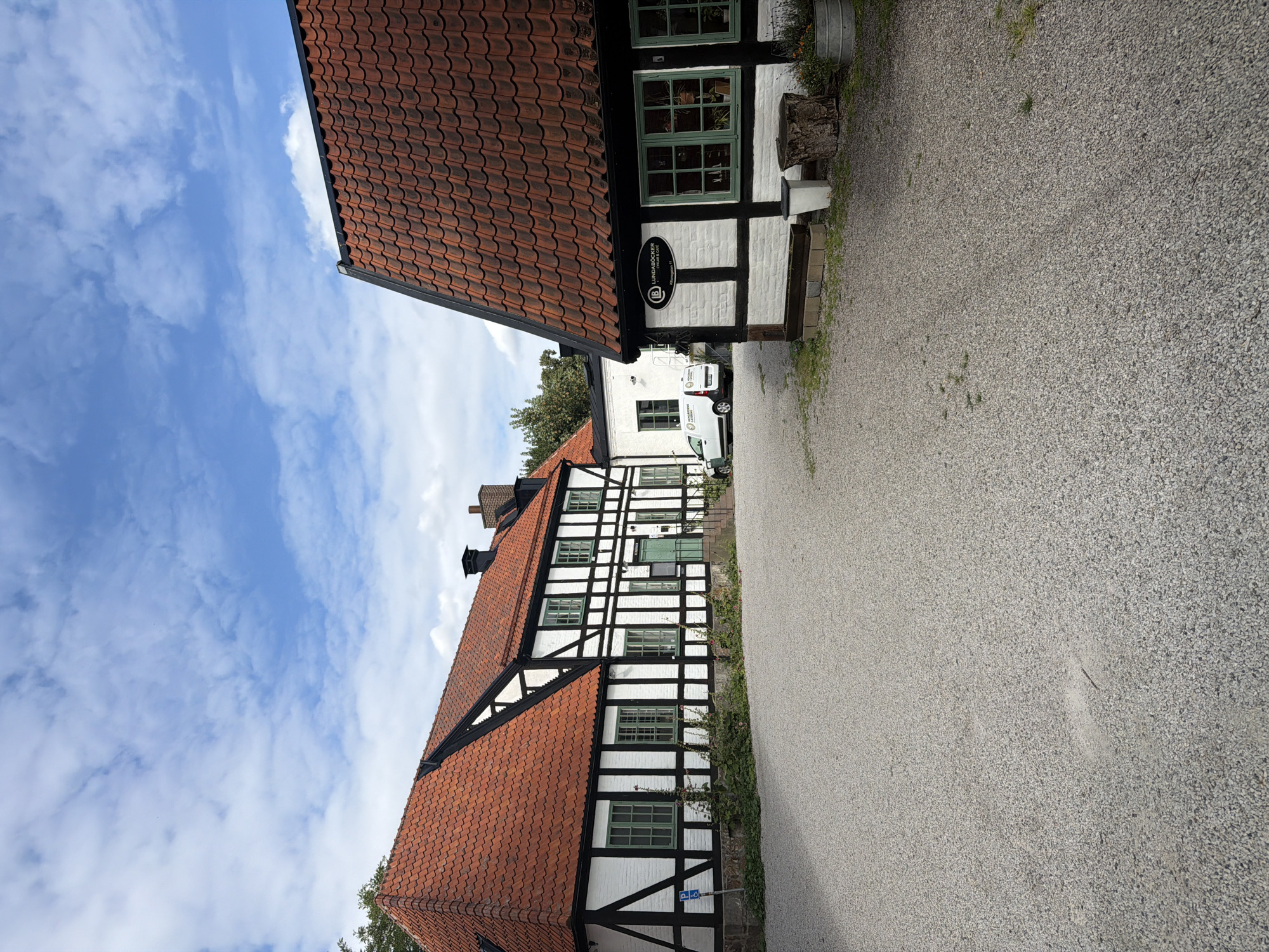 A wide-angle, eye-level shot depicts a gravel courtyard in front of a white and black timber-framed building with a red tile roof. The building features green-paned windows. To the right, another similar building is partially visible, also with a red tile roof and green window panes. A white van with "VÄRNAMO" text is parked in the background. The sky above is blue with scattered white clouds. The overall scene suggests a historical or rural setting. The building on the right has a sign that reads "LUNDABÖCKER CYKLAR & CAFÉ" and "Källängsvägen 11". This indicates the location is Lundaböcker, a shop and cafe specializing in books, bicycles, and coffee, situated at Källängsvägen 11 in Lund, Sweden.