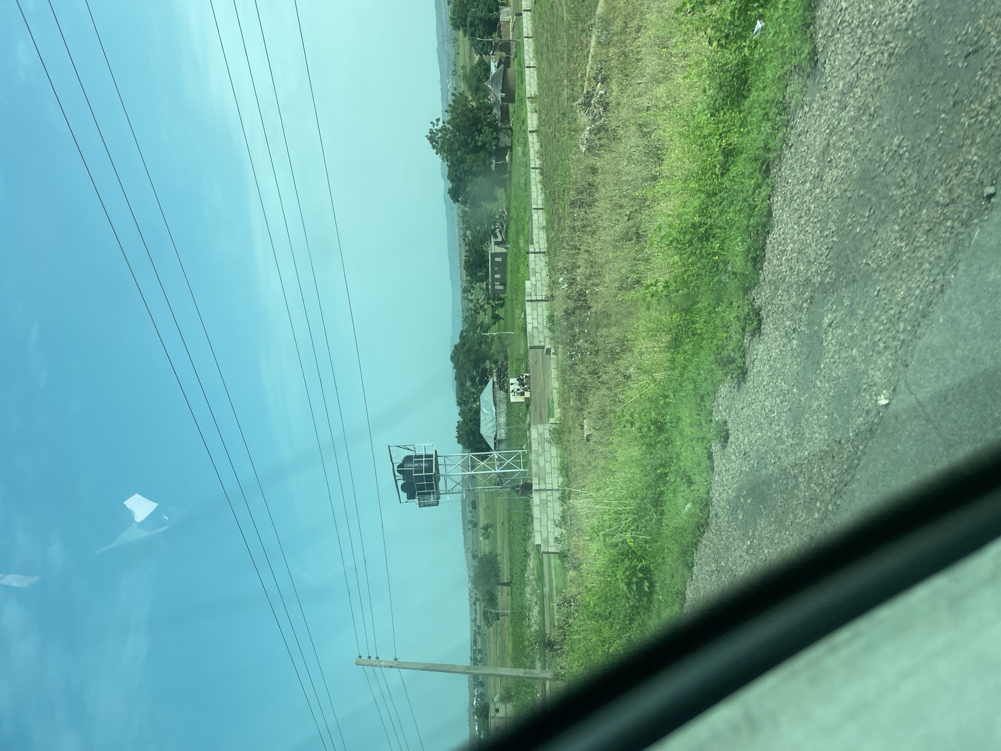 The image shows a view from a vehicle, looking out at a rural landscape in Chum, Nigeria. In the foreground, a gravel road is visible, partially obscured by the vehicle's window frame. Beyond the road, there is a field of tall green grass and some dry brush. Further back, a prominent structure is a tall metal water tower with several black water tanks on top. A single utility pole stands to the left of the water tower, with power lines stretching across the sky. In the middle ground, there are several buildings, including what appear to be small houses with thatched roofs and some brick structures. A low brick wall runs across part of the landscape. In the distance, rolling hills and trees can be seen under a bright, clear blue sky with some faint clouds. There are a few people visible near the buildings in the middle ground. The overall scene depicts a sparsely populated rural area with evidence of infrastructure and dwellings.