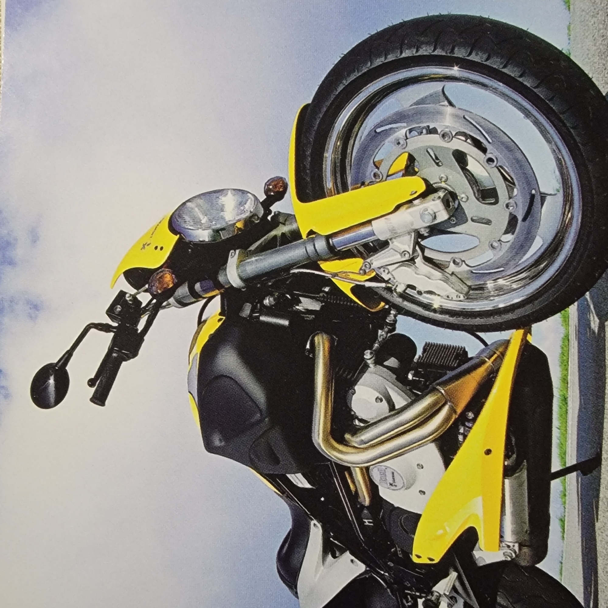 The image is a low-angle, close-up shot of a yellow and black motorcycle parked on an asphalt surface. The motorcycle is the primary subject, with its front tire and lower half prominently featured. The background consists of a hazy blue sky with some clouds. The motorcycle appears to be in good condition, with a shiny chrome exhaust pipe and a reflective chrome wheel rim. The yellow fairing is a vibrant accent against the darker body of the bike. There are no people visible in the image. The scene is static, with the motorcycle appearing to be stationary. The lighting suggests it could be daytime, possibly overcast, given the softness of the shadows.