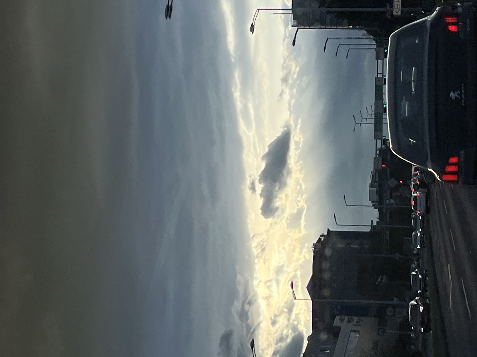 The image depicts a street scene in what appears to be a densely populated area of City of Zagreb, Croatia. The foreground is dominated by the rear of a dark-colored car with visible taillights. Other vehicles are visible in the lane. On the left, a cloudy sky appears with a distinct break in the cloud cover, allowing sunlight to filter through. The streetscape includes tall buildings with visible windows and rooftop structures, interspersed with streetlights. Traffic signals and signs are also present along the street. The image is taken from a relatively low angle, presumably from within a vehicle.