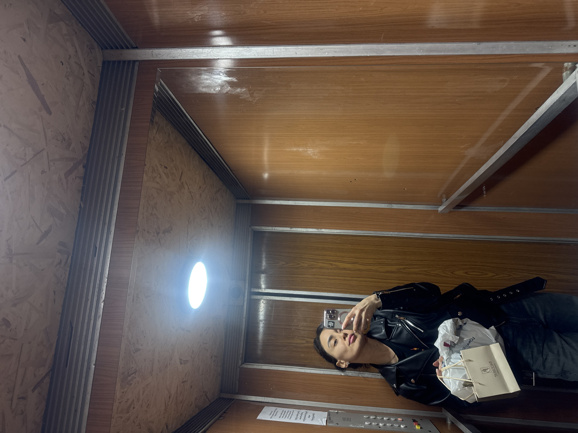 A woman is taking a selfie in an elevator. The elevator has light wood paneling on the sides and ceiling, with a circular light fixture on one side. The woman is wearing a black leather jacket and holding a shopping bag. There are elevator buttons visible at the bottom of the frame. The Cyrillic writing on the shopping bag identifies it as "Tavamsagskaia." Based on this information, it is possible that the location is in Tbilisi, Georgia.