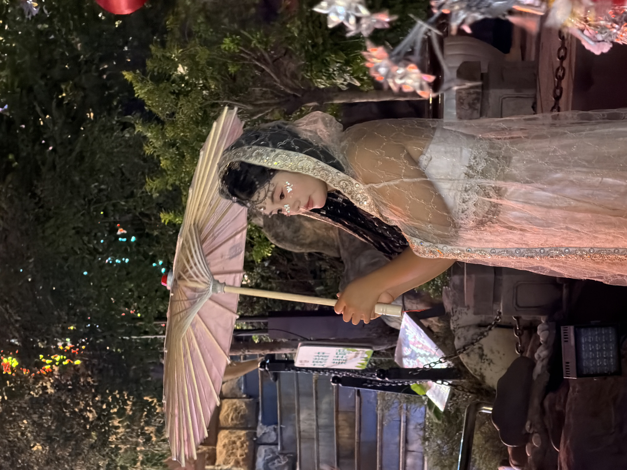 A young East Asian female, dressed in a white gown and a sheer, ornate veil with beaded trim, stands outdoors at night, holding a light pink parasol. She has glitter applied to her face and her dark hair is styled in braids. The background features lush green trees illuminated by colorful, blurred lights. To the left, a stone staircase with a black metal railing leads upwards. A sign with Chinese characters is visible on a post near the stairs. The ground is covered in rocks and a small, dark electronic device with a grid of lights sits on the lower left. The overall setting appears to be an outdoor cultural or recreational space, possibly within Yuzhong District, China, given the context.