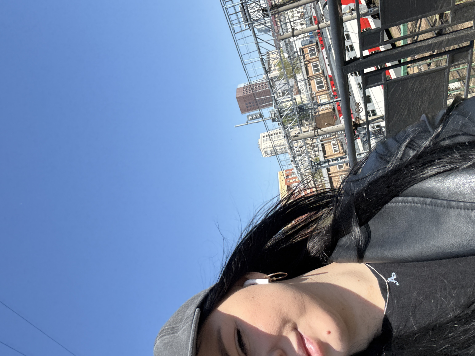The image shows a person wearing a gray hat, a black leather jacket and a black top with a small silver pendant. They have black hair, and are wearing an earbud. The background includes buildings, possibly a cityscape in Moscow, Russia. There is also a metal structure or railing visible. The sky is clear and blue. The camera appears to be held at an unusual angle.