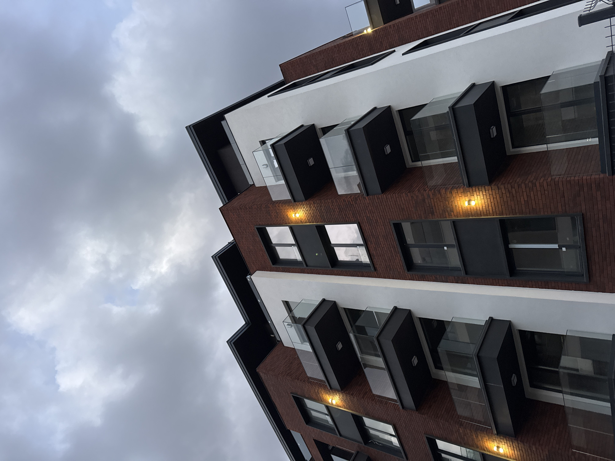 A low-angle shot captures a modern building against a cloudy sky, possibly located in Sofia, Bulgaria. The building features a brick facade with white horizontal sections. The windows are framed by black, rectangular structures that project outwards, creating a unique architectural detail. Some of these structures are enclosed with transparent glass. The sky is overcast, with grey clouds dominating the upper portion of the frame. There are no people visible in the image.