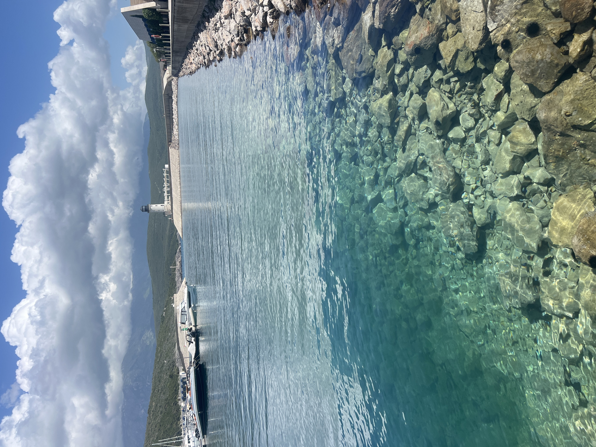 A clear, calm body of water borders a rocky shoreline and a concrete structure, with glimpses of boats and a lighthouse in the distance. The clarity of the water allows visibility of rocks beneath the surface. The scene is located in Radovići, Montenegro. Cumulus clouds are present.
