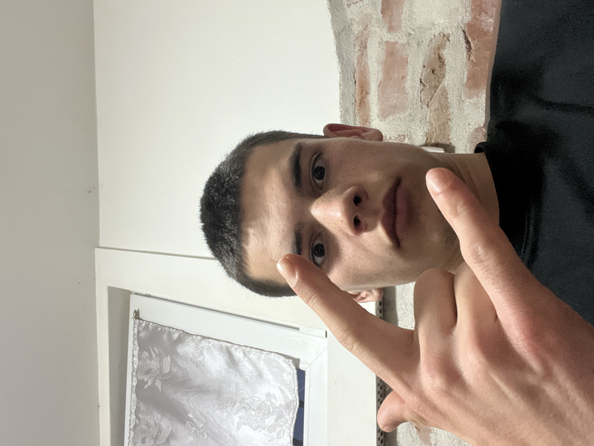 A young man is facing the camera and gesturing with his hand. The background includes a white wall, a window with a white curtain, and a brick wall. There is no evidence indicating the location to be Plovdiv, Bulgaria.