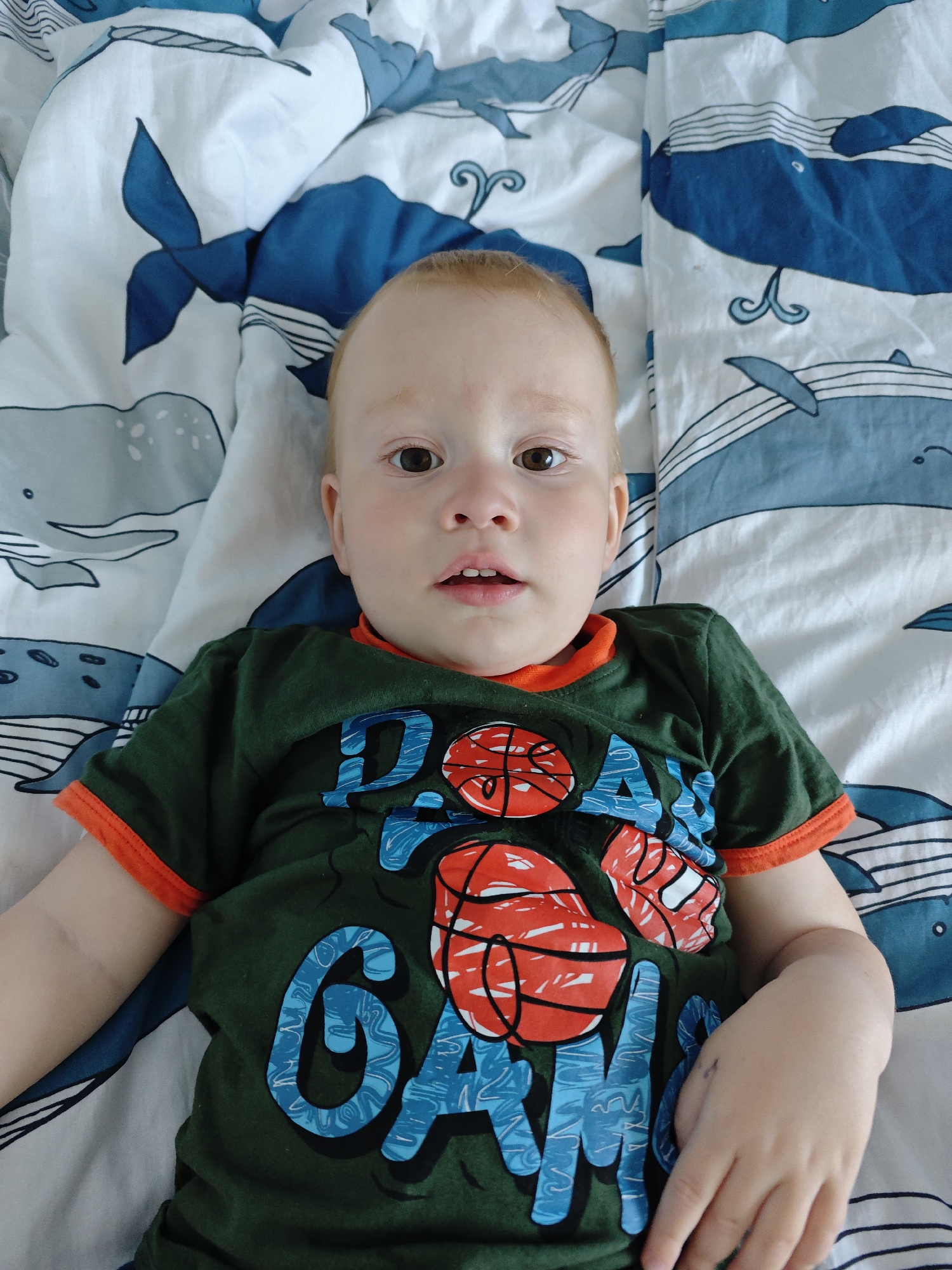 A toddler is lying on a bedspread with a blue and white whale pattern. He is wearing a green t-shirt with orange trim and a blue and red basketball design. The toddler is looking directly at the camera with his mouth slightly open. The location is indeterminate, though the user specifies it as Markovskiy, Russia.