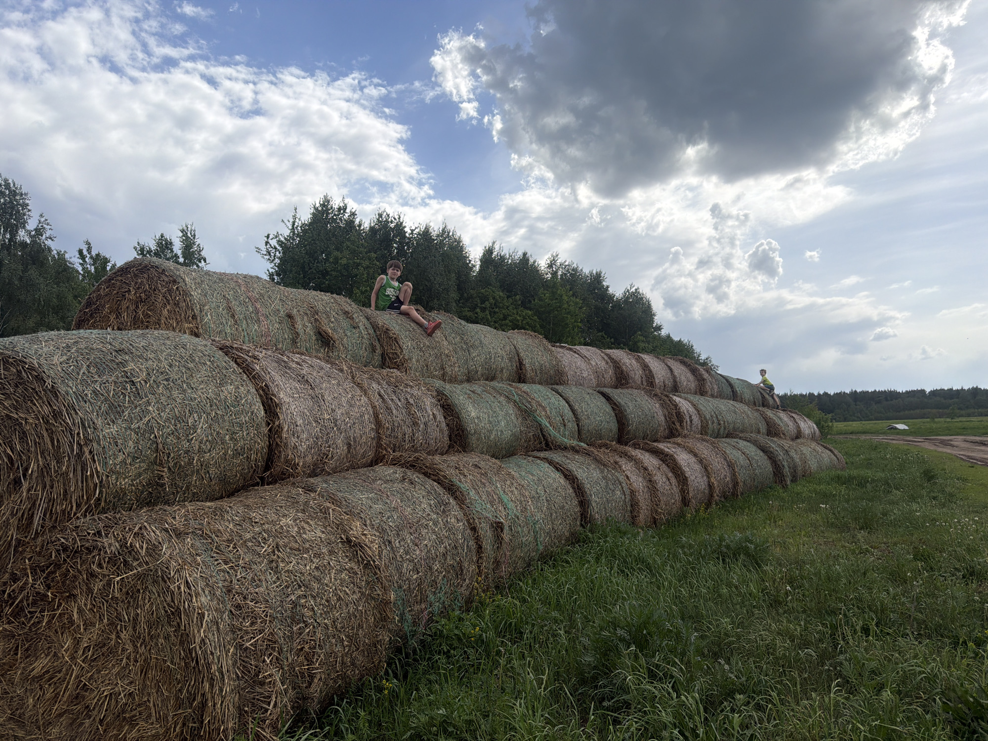 The image shows a stack of hay bales in a field. There are two people visible on the stack. One person is on the top of the stack and is wearing a green shirt and red shoes. Another person is on the top right of the stack. The field is grassy with some dirt in the background. The sky is cloudy. The image was taken in Kirillovka, Russia.