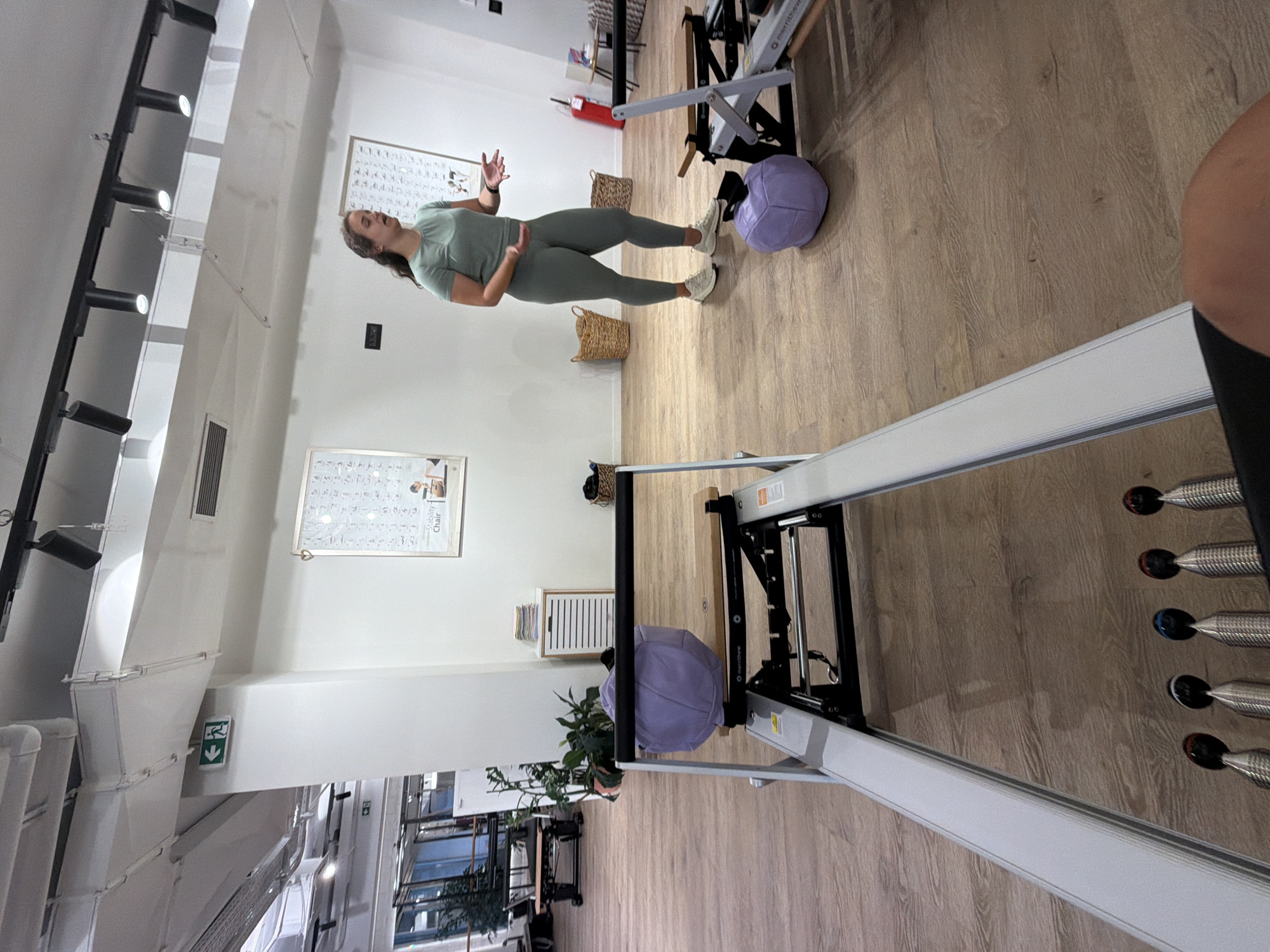 The image depicts an indoor Pilates studio with wooden floors and white walls. In the foreground, a piece of Pilates equipment, likely a reformer, is partially visible. In the mid-ground, a woman wearing a green athletic outfit stands with her hands gesturing as if explaining something. Two large, purple exercise balls are present on the floor. In the background, there are framed charts on the wall, plants, storage units, and other fitness equipment. An emergency exit sign is visible on the wall. The scene is brightly lit by overhead lights. The location is identified as Boreti, Montenegro.