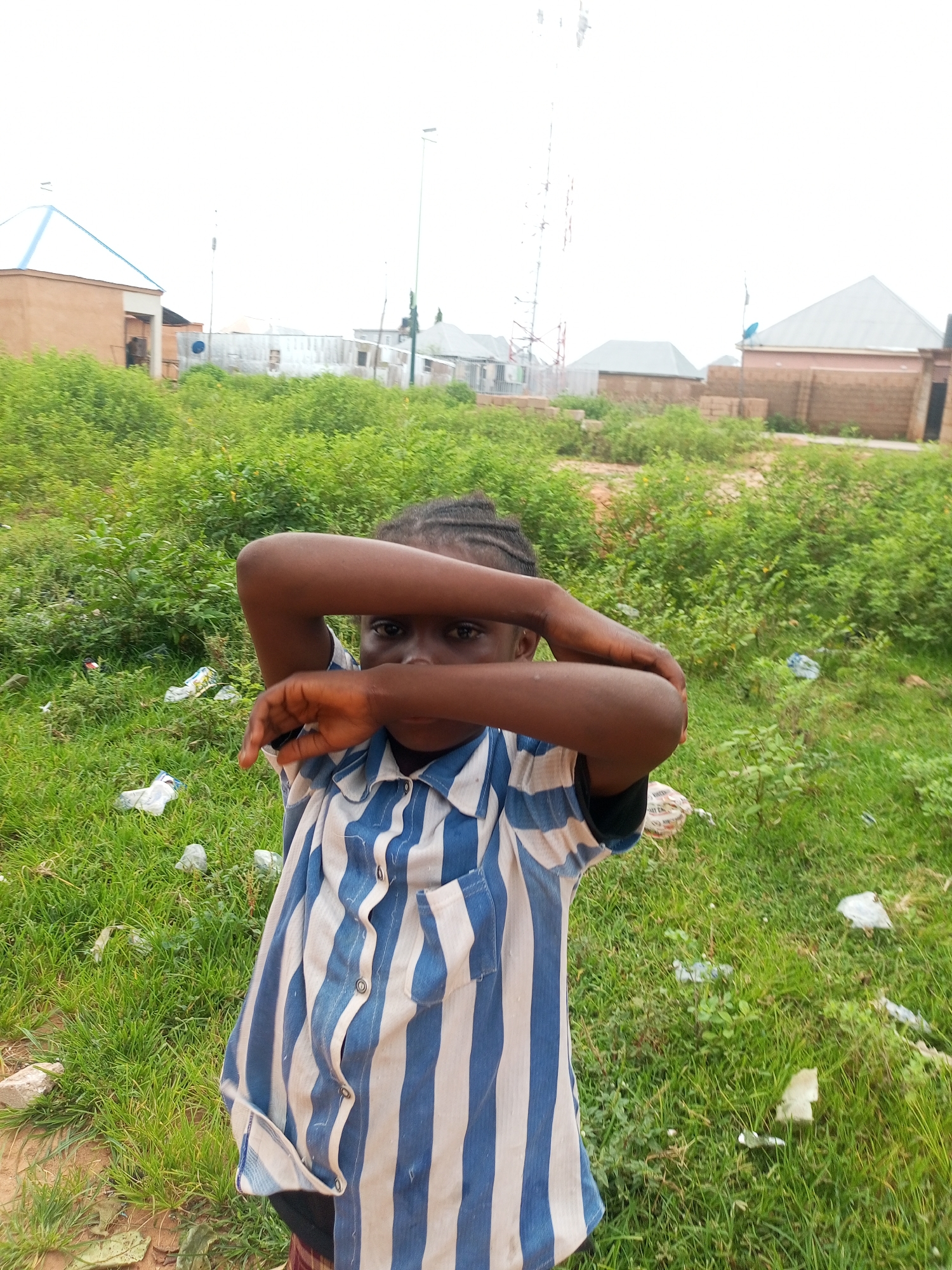 A young girl with dark skin and braided hair stands in a grassy, overgrown area. She is wearing a blue and white striped shirt. Her arms are crossed in front of her face, obscuring it. In the background, there are several buildings with corrugated metal roofs and brick walls, and what appears to be a cell tower. The ground is covered in green grass and scattered debris, including plastic bottles and paper. The sky is overcast and white. The scene is outdoors, likely in a developing urban area. This image is taken in Tempure, Nigeria.