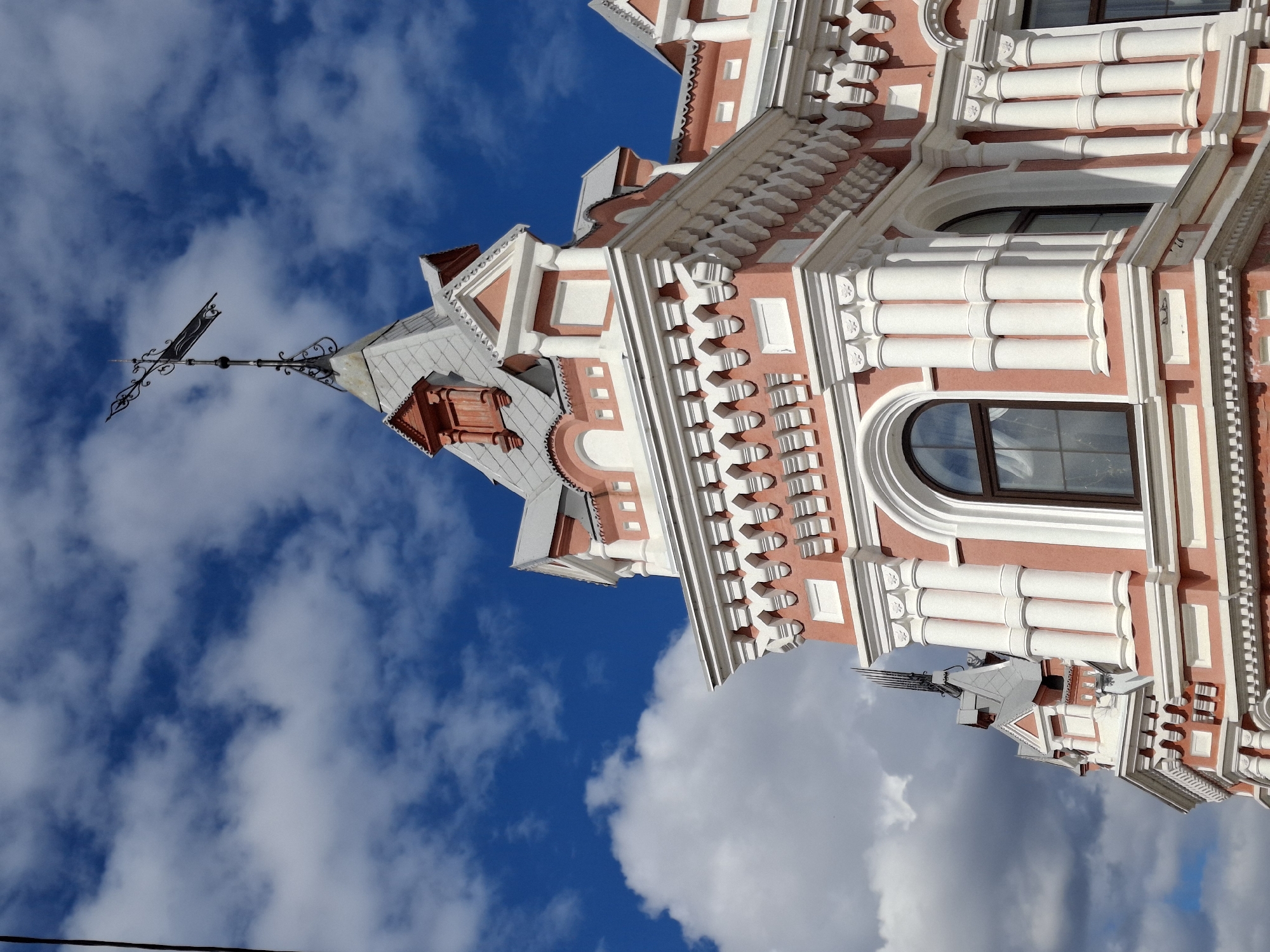 The image displays the ornate upper section of a historical building against a vibrant blue sky dotted with scattered white clouds, characteristic of daytime. Located in Sarapul, Russia, the architecture is distinguished by its salmon-pink walls richly contrasted with an abundance of white decorative elements.
The building's facade features intricate detailing, including stacked columns or pilasters, elaborate cornices, balustrade-like motifs, and patterned friezes that create a highly textured and decorative appearance. Several arched windows, framed in dark wood or metal, are visible, reflecting the bright sky. Shadows cast by the architectural projections indicate strong, clear sunlight.
Towards the upper left, a prominent spire or turret rises, topped with a greyish roof adorned with small red-brown accents. Crowning this spire is a striking black, ornate weathervane, shaped like a decorative arrow with intricate scrollwork, standing sharply against the azure backdrop.
The sky is a dominant element, a brilliant blue with fluffy, irregularly shaped white clouds suggesting fair weather. There are no people visible, and no specific activities or events are depicted, making it a static architectural study. A thin black wire is barely visible at the very bottom edge of the original image. No visible text is present on the building or in the scene.