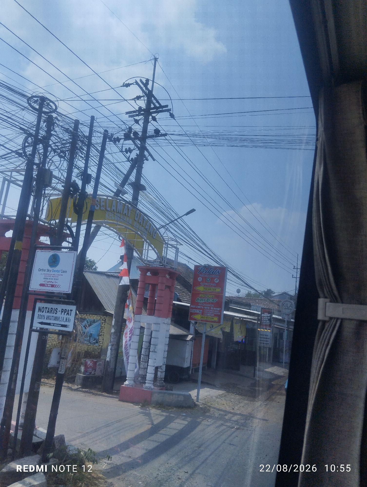 The image depicts a street view in Kraton, Indonesia, with a prominent archway bearing the Indonesian greeting "SELAMAT DATANG" (Welcome). The archway is adorned with numerous overhead electrical wires and poles, creating a dense network against a bright blue sky with scattered clouds. Various signs are visible along the street, including one for "Ortho Sky Dental Care" and another for "NOTARIS - PPAT RIZKYA ANGGITAWAN, S.H., M.Kn." Further down the street, more signs are visible, including one for "Pedals" which appears to be a food establishment, and another for "KILA bakery." The street itself is paved, and there are buildings and structures on either side. The overall impression is of a commercial or residential area within an Indonesian town, characterized by its overhead infrastructure and signage. The timestamp on the image indicates the date August 22, 2026, at 10:55 AM.