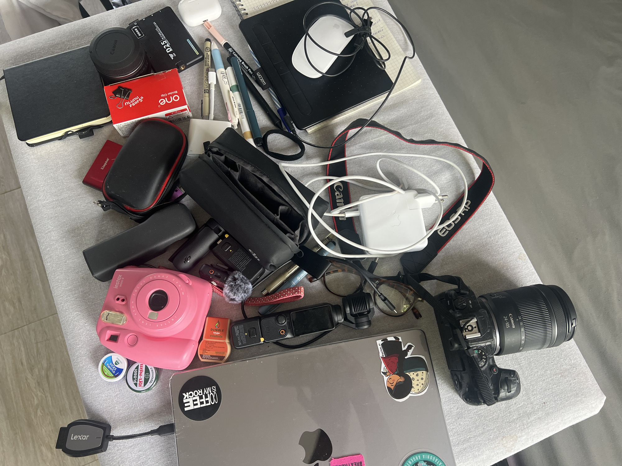 The image shows a tabletop cluttered with various objects. Notable items include a Canon camera with a 24-105mm lens, a pink Instax Mini 9 camera, a laptop with stickers including one that reads "Coffee is my rock," a Wacom tablet, a white computer mouse, AirPods in their case, notebooks, pens, a DJI Osmo Pocket with a microphone, glasses, various cases for electronics, a lens cap, a box of binder clips, a power adapter with associated cables, and small containers of Vicks VapoRub and Dumble Inhaler. A Lexar memory card reader is connected to the laptop. The environment consists of a gray fabric-covered surface. Location information from the objects (stickers, products) suggests a possible connection to Thailand, specifically บ้านบางรักษ์.